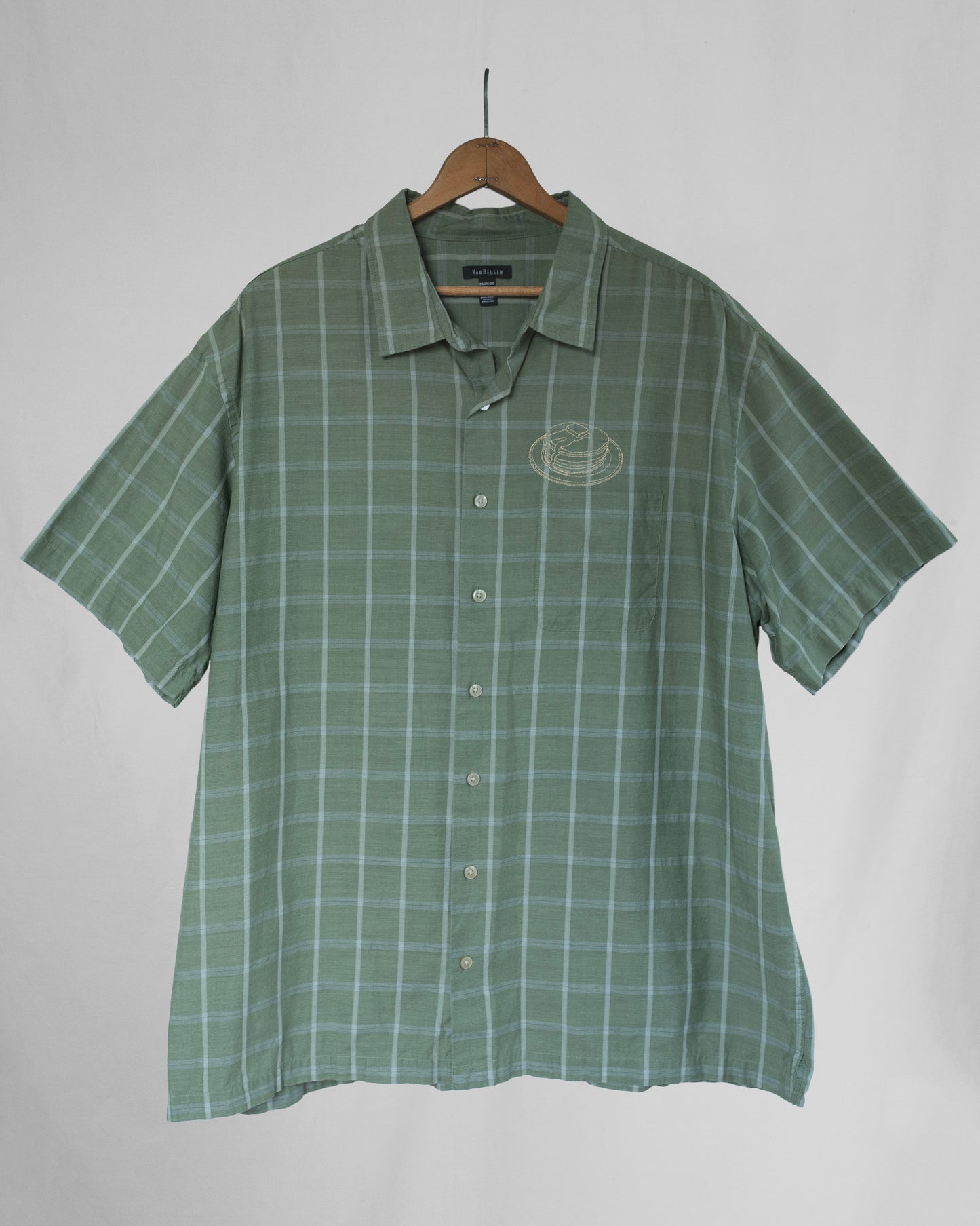 Green checkered shirt with pancakes embroidered above the chest pocket on a hanger against a plain background