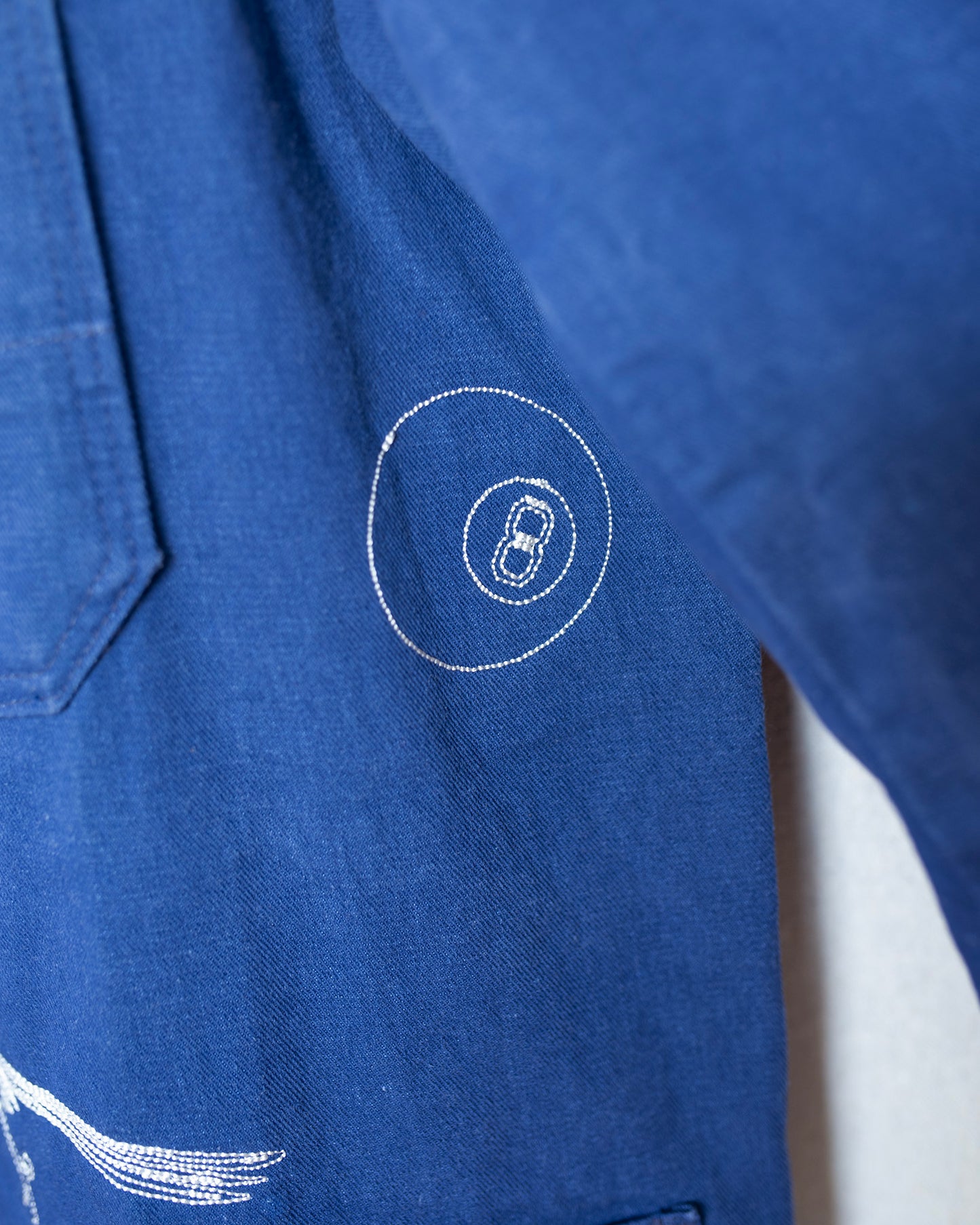 Close-up of a blue canvas garment with white embroidered details, including a circular stitched motif and part of a wing-like design.