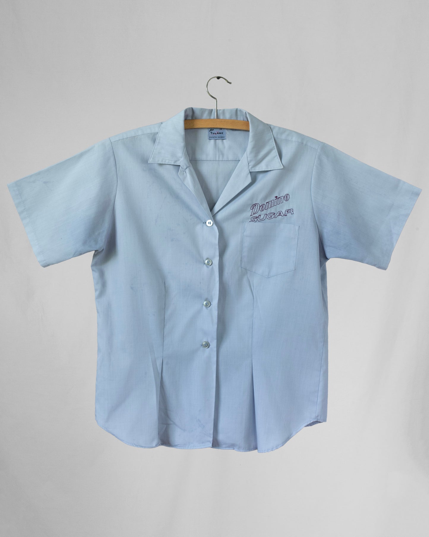 Light blue short-sleeve shirt with embroidery above chest pocket on a hanger against a gray background