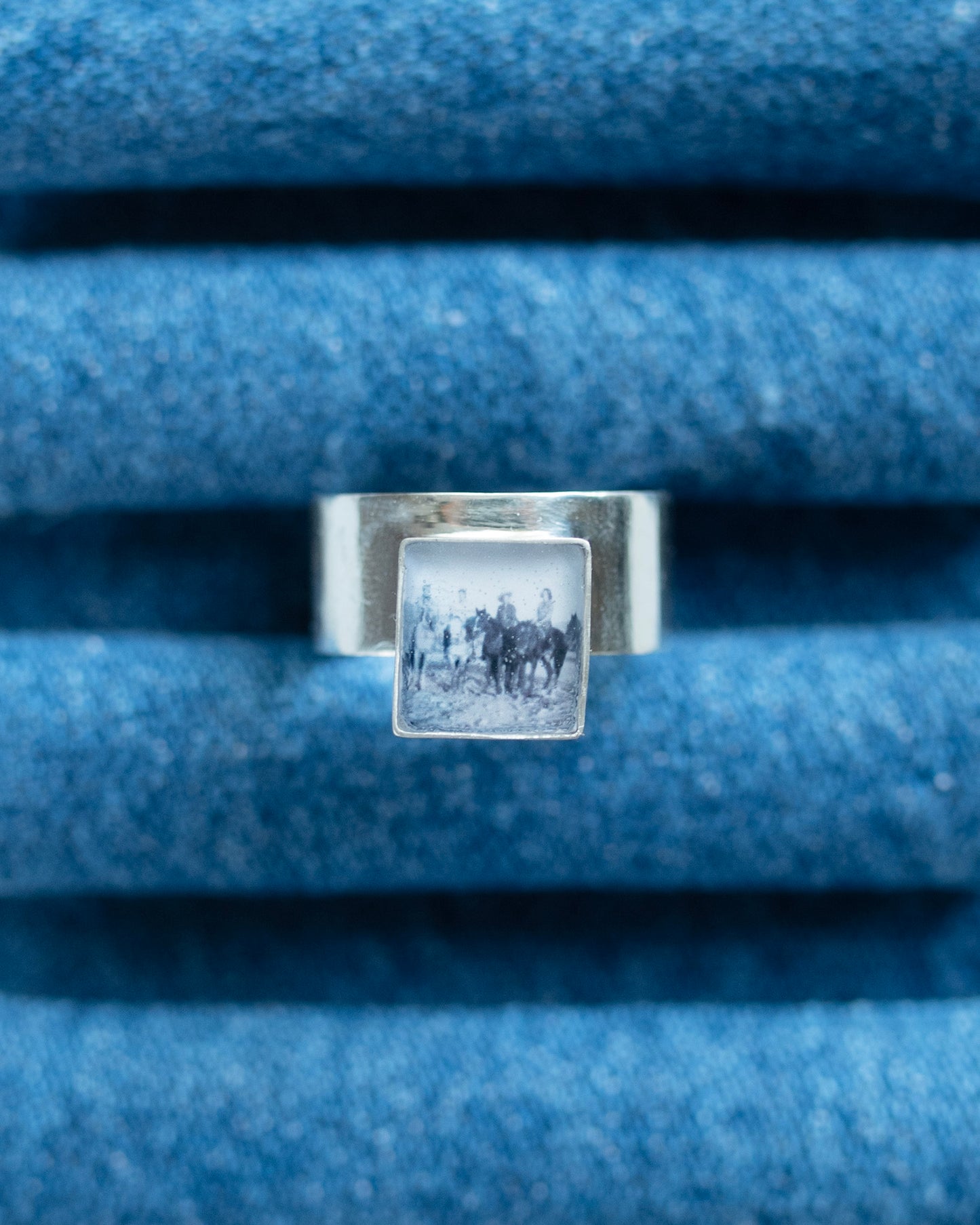 A silver ring with a square bezel containing a black-and-white vintage photograph of a horse riders, displayed on a textured blue denim ring stand.