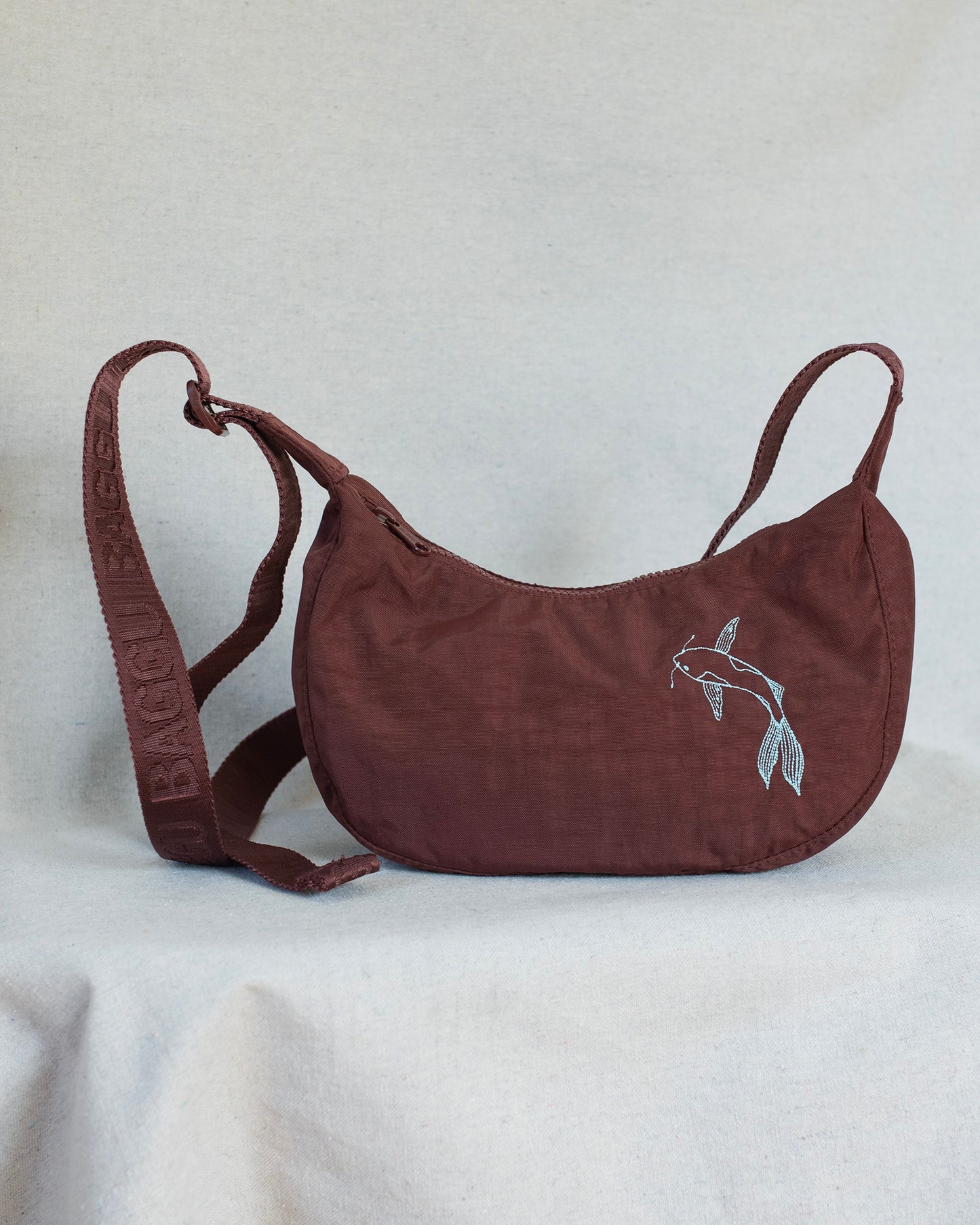 A mahogany crescent-shaped shoulder bag with a matching adjustable strap, featuring a white embroidered koi fish design on the front and 'BAGGU' embossed on the strap, set against a light gray fabric backdrop.