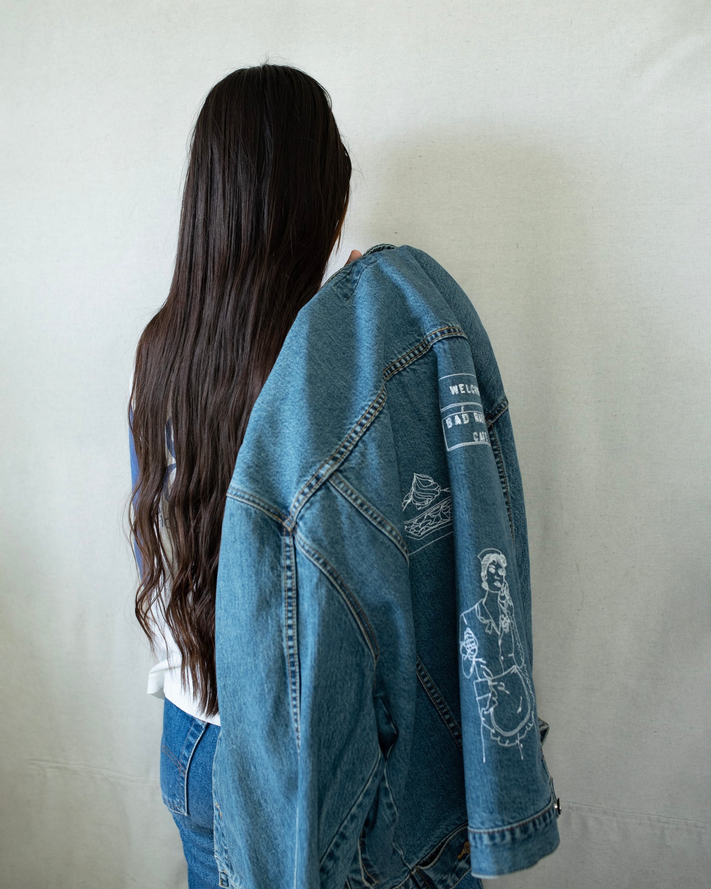 The back of a girl holding a denim jacket over her shoulder, showing the embroideries on the back of it.