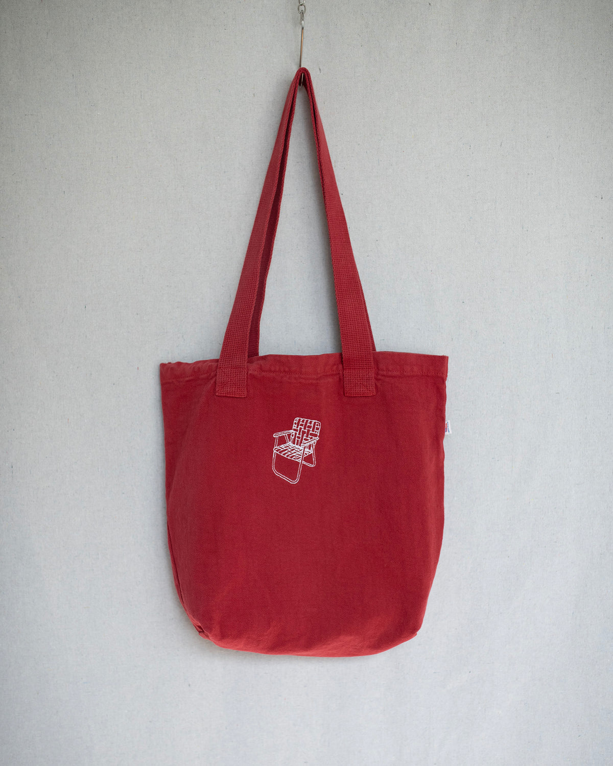 Hanging red canvas tote bag that has a small lawn chair embroidery on top center.