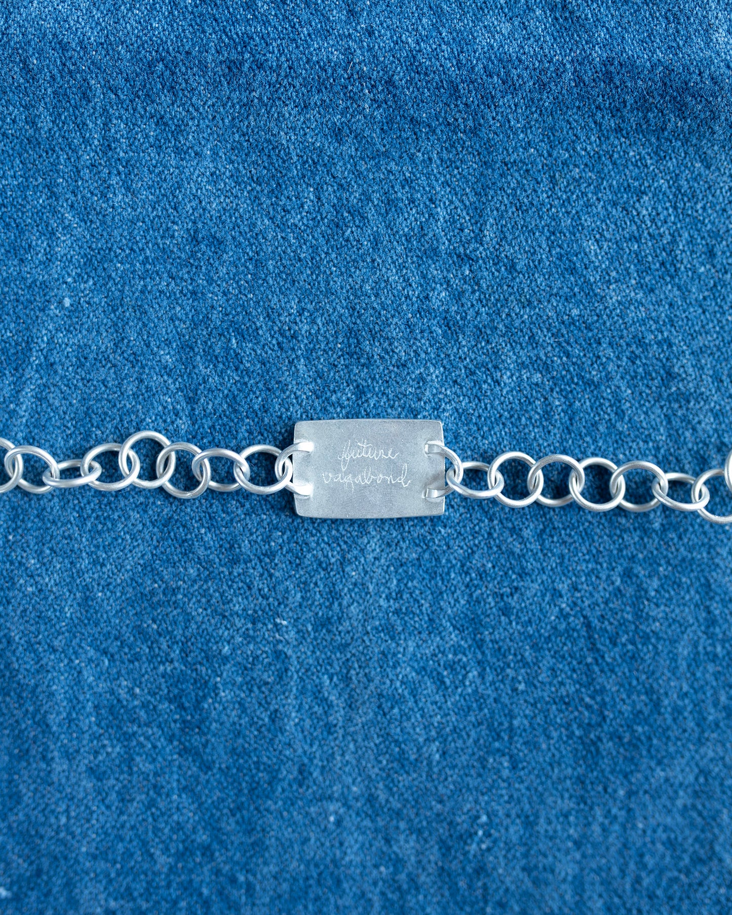 A silver chain bracelet with a rectangular engraved nameplate, laid flat on a textured blue fabric background.