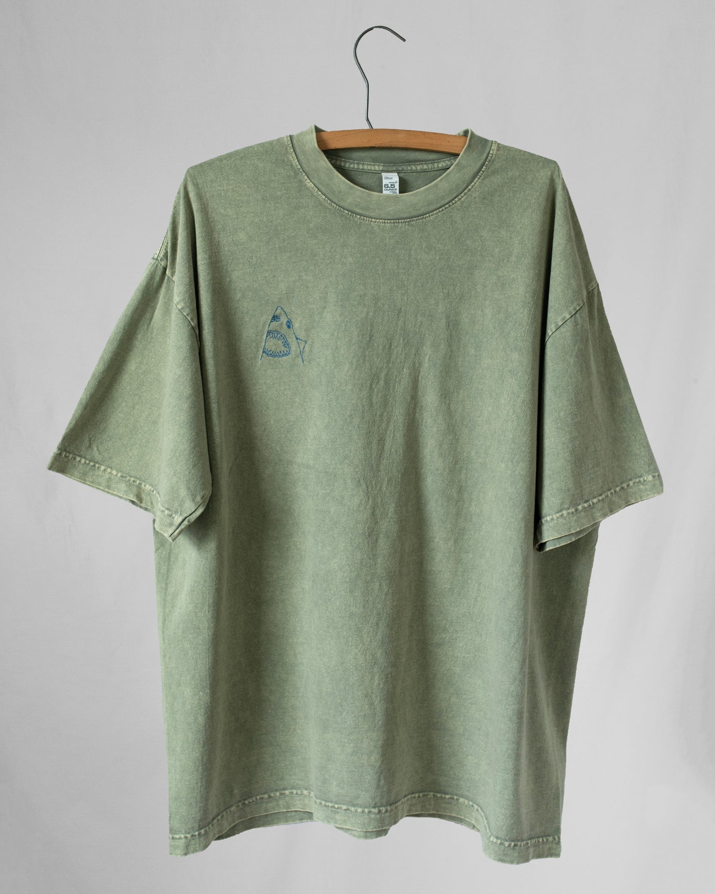 A green, crew neck t-shirt with an embroidered shark on the chest, displayed on a hanger against a white background.