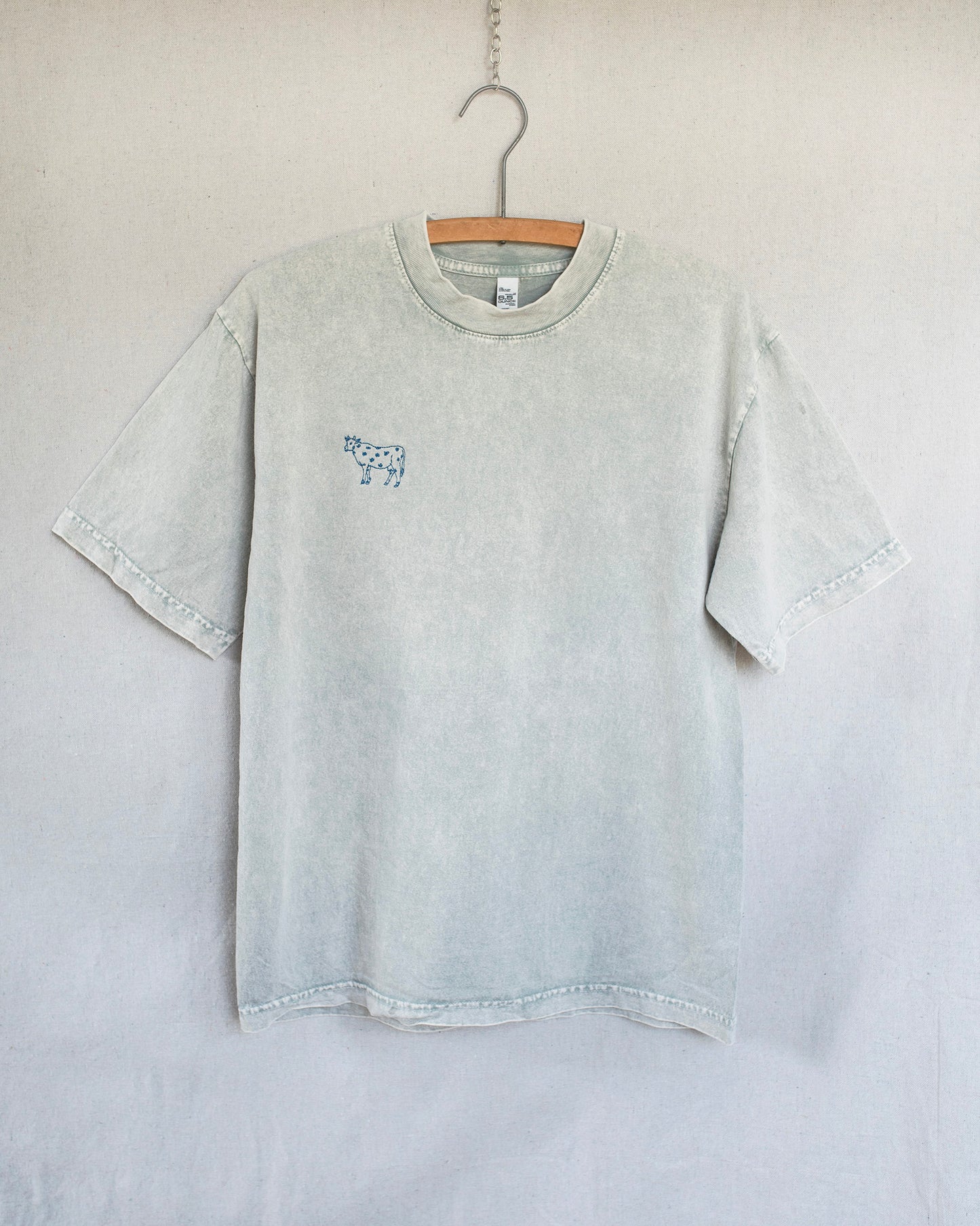 A light gray mineral-wash short-sleeve t-shirt hanging on a wooden hanger against a neutral light gray wall. The shirt features a small blue graphic print on the left chest depicting a cow..