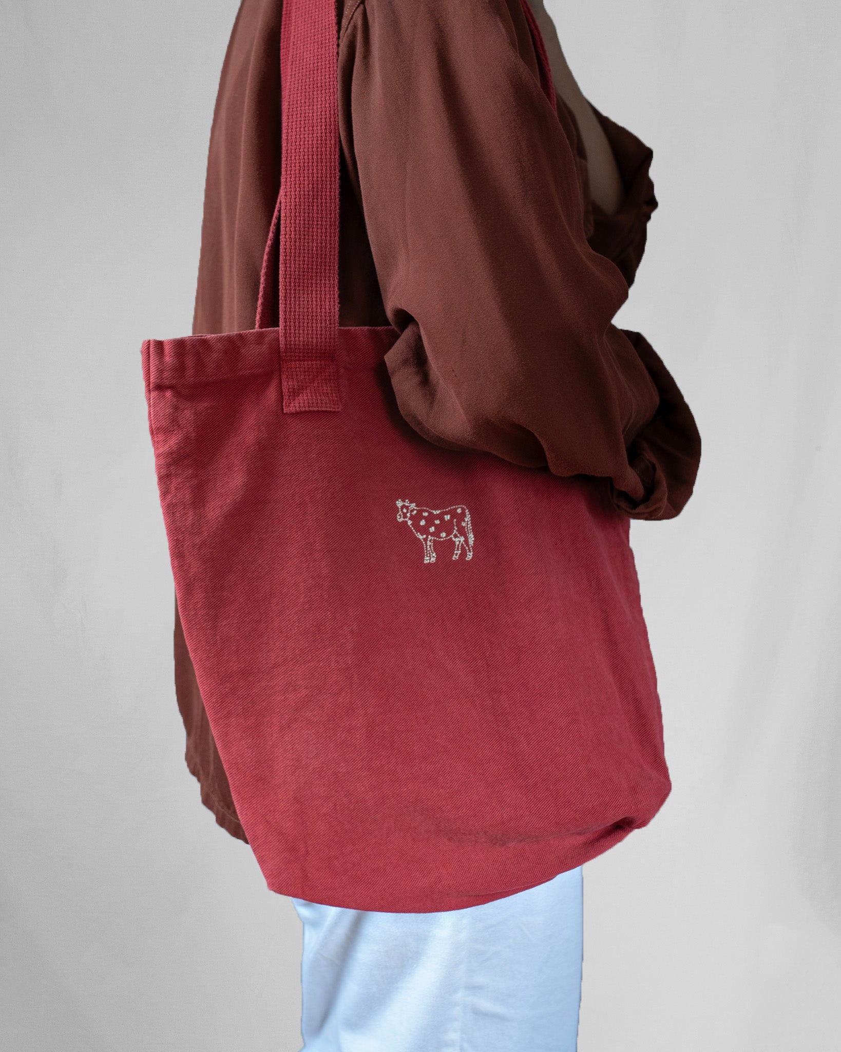 Red tote bag with a small logo held by a person wearing a brown jacket and light blue pants on a plain background.