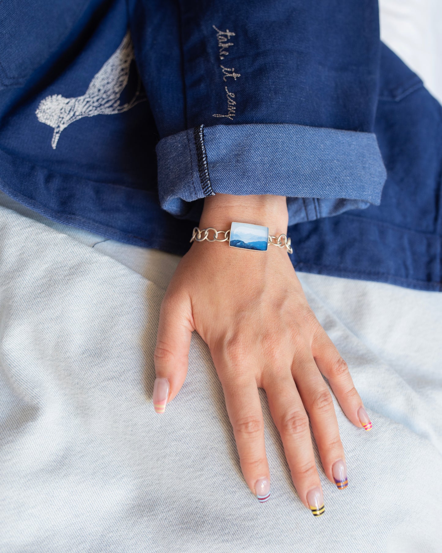 Close up of a hand wearing a silver bracelet with mountains charm set behind light blue denim and a darker blue embroidered jacket.