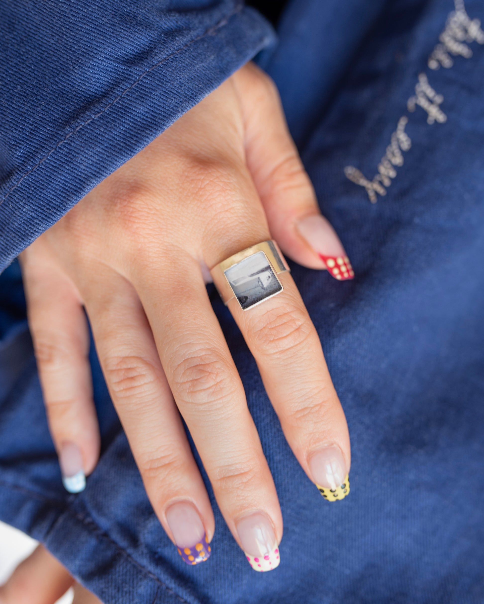 A close-up of a hand wearing a large sterling silver square ring with a black-and-white photograph mounted in the center, resting on blue canvas fabric.