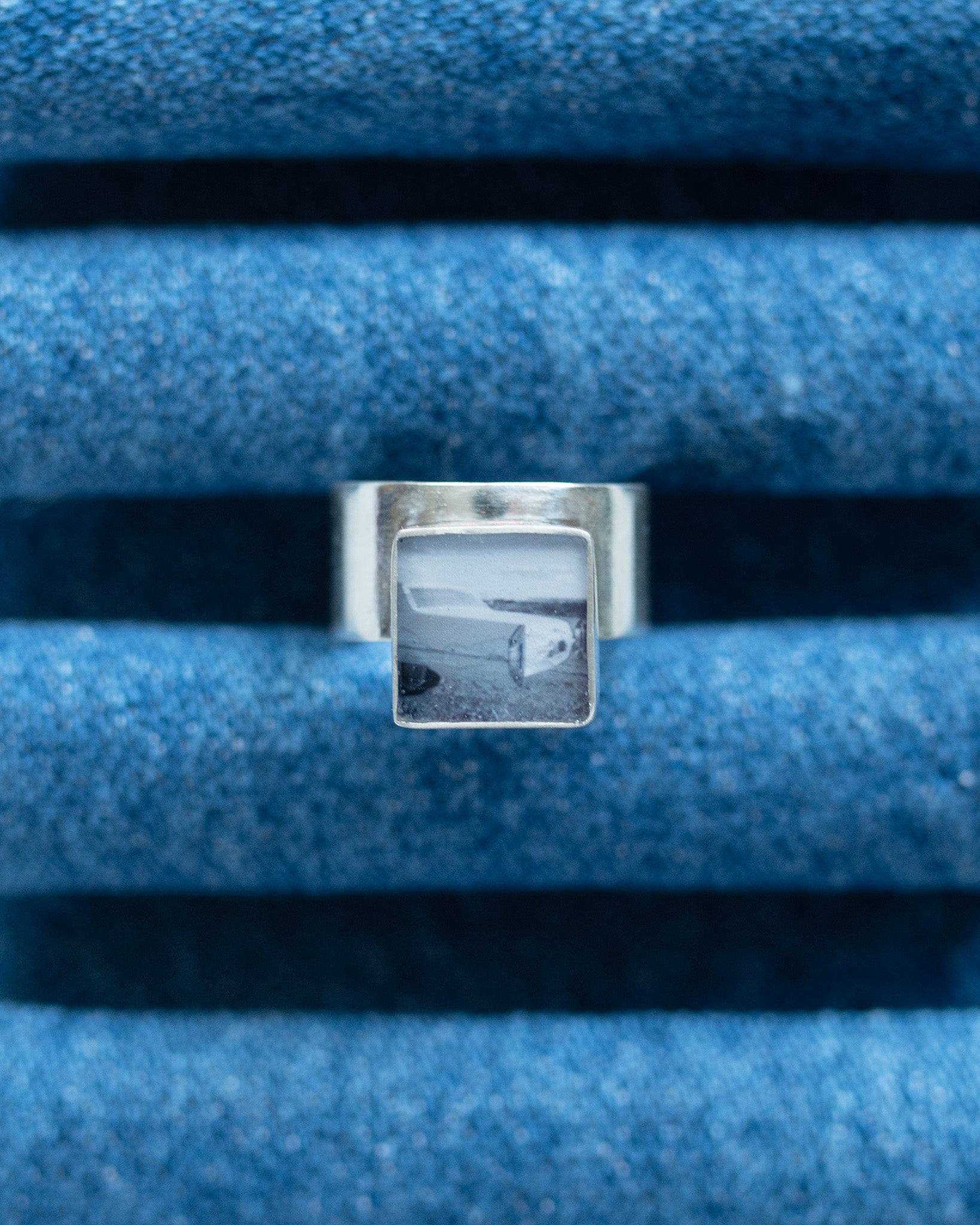 A silver ring with a square bezel containing a black-and-white landscape photograph, displayed on a textured blue dennim ring stand.