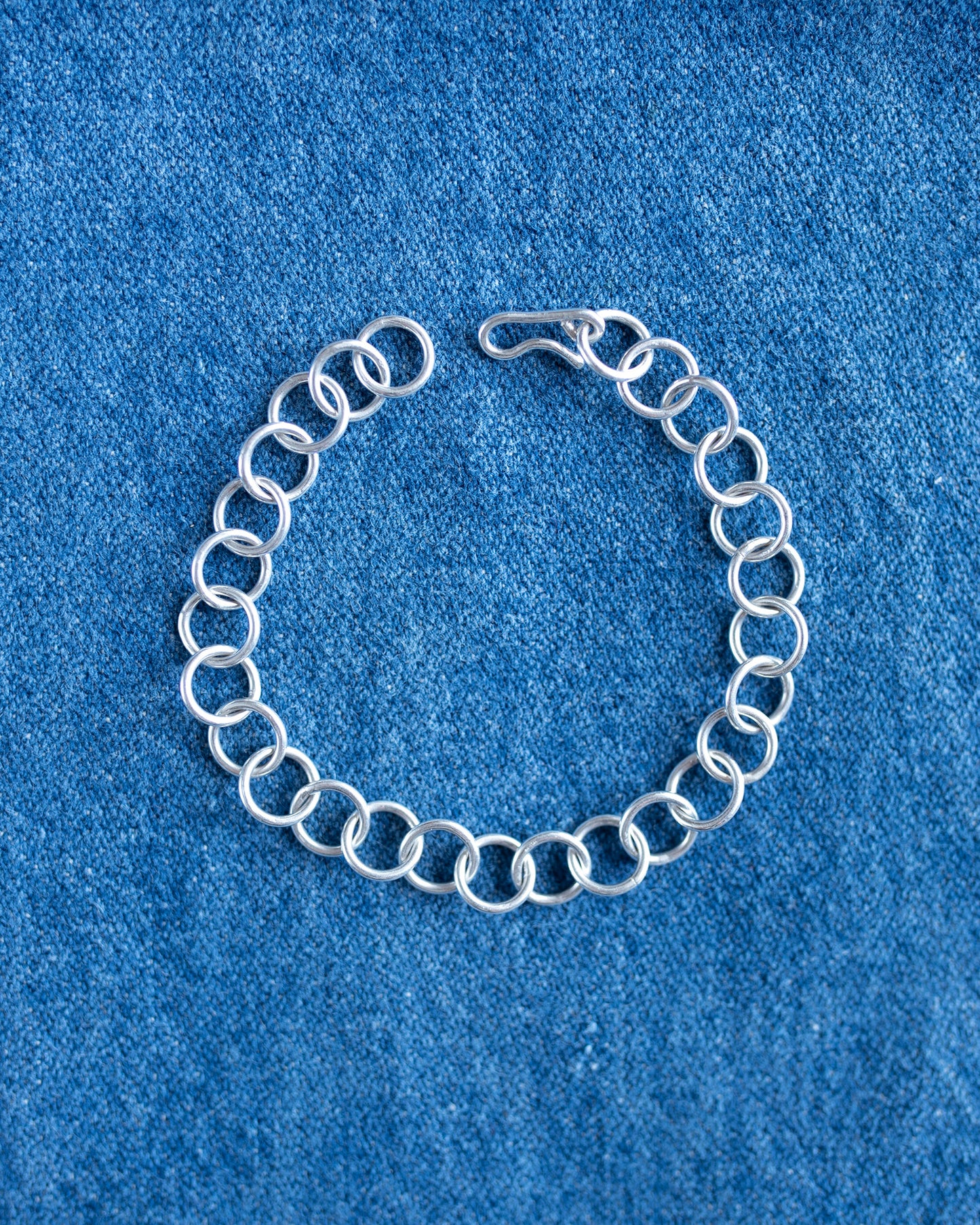 A silver chain link bracelet with a spring-ring design and a lobster clasp, displayed on a textured blue fabric background.