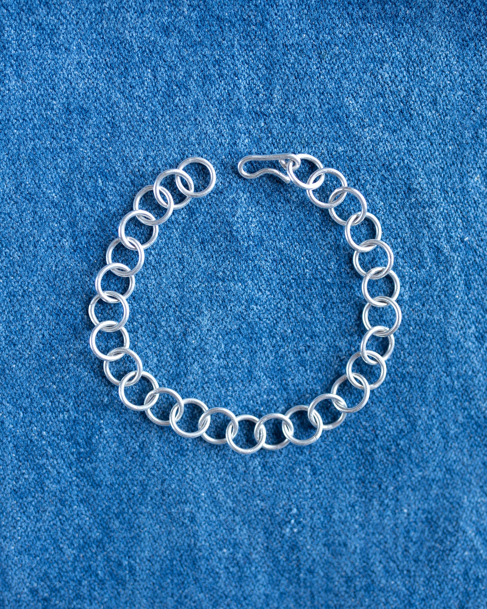 A silver chain link bracelet with a spring-ring design and a lobster clasp, displayed on a textured blue fabric background.