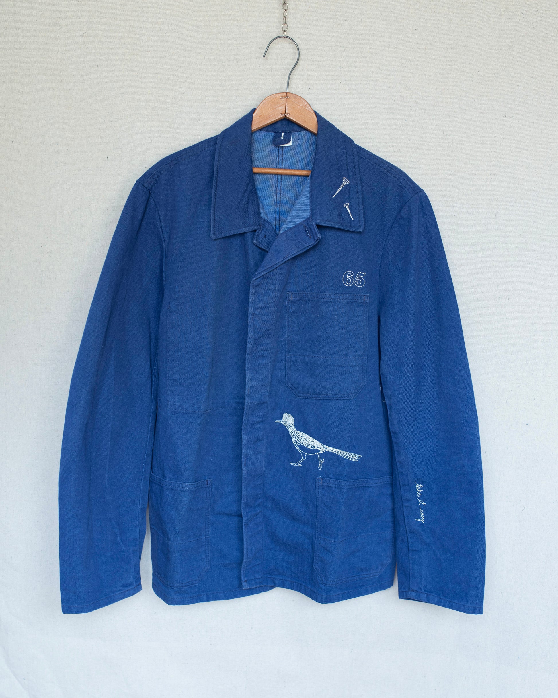 A blue indigo work jacket hanging on a wooden hanger against a light gray wall. The jacket features white embroidered details, including a bird motif above the front pocket and a small 'G5' logo on the chest pocket.