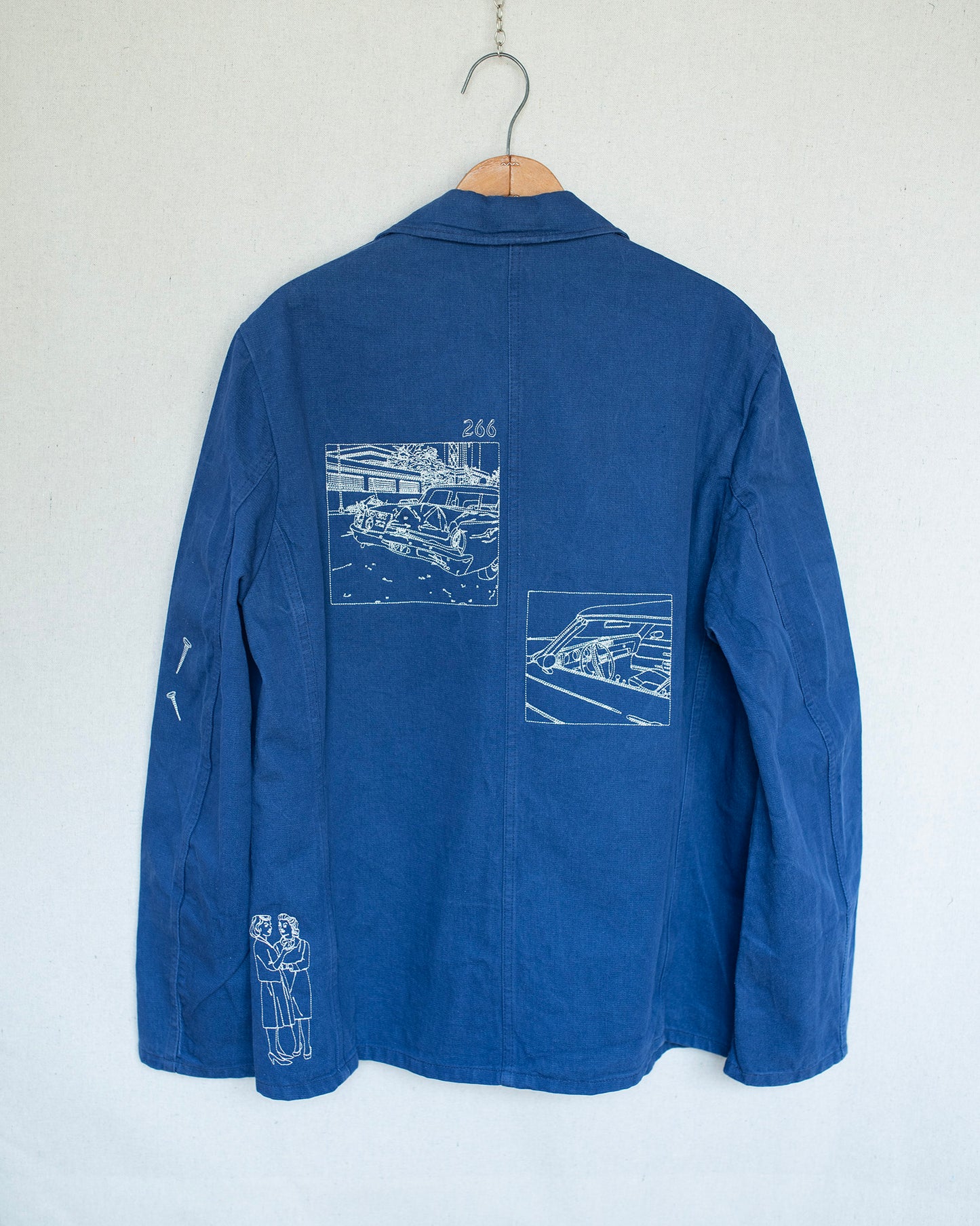 A blue jacket back hanging on a wooden hanger against a white wall, featuring white line-art illustrations of cars and a small graphic on the back and left sleeve.