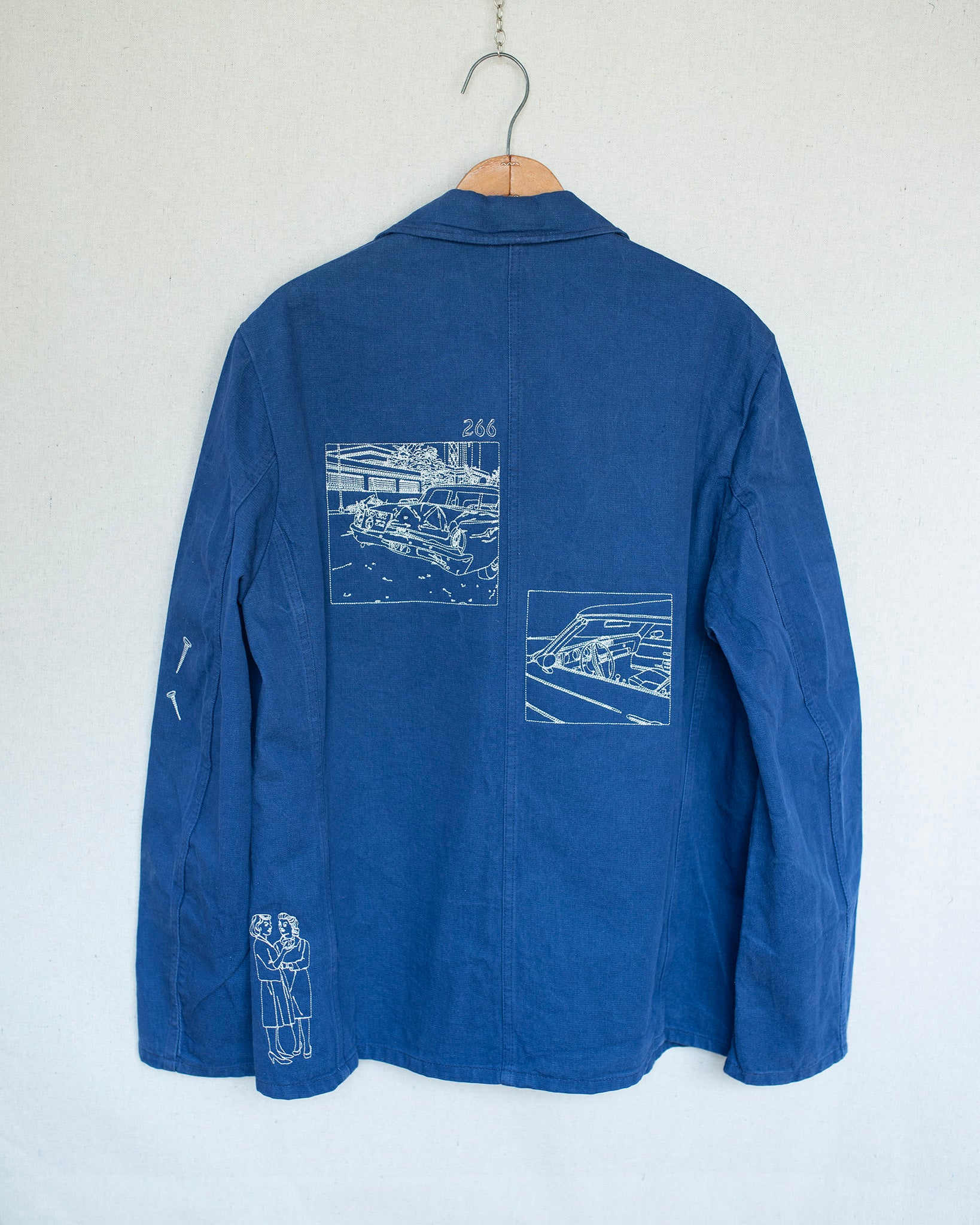 A blue jacket back hanging on a wooden hanger against a white wall, featuring white line-art illustrations of cars and a small graphic on the back and left sleeve.