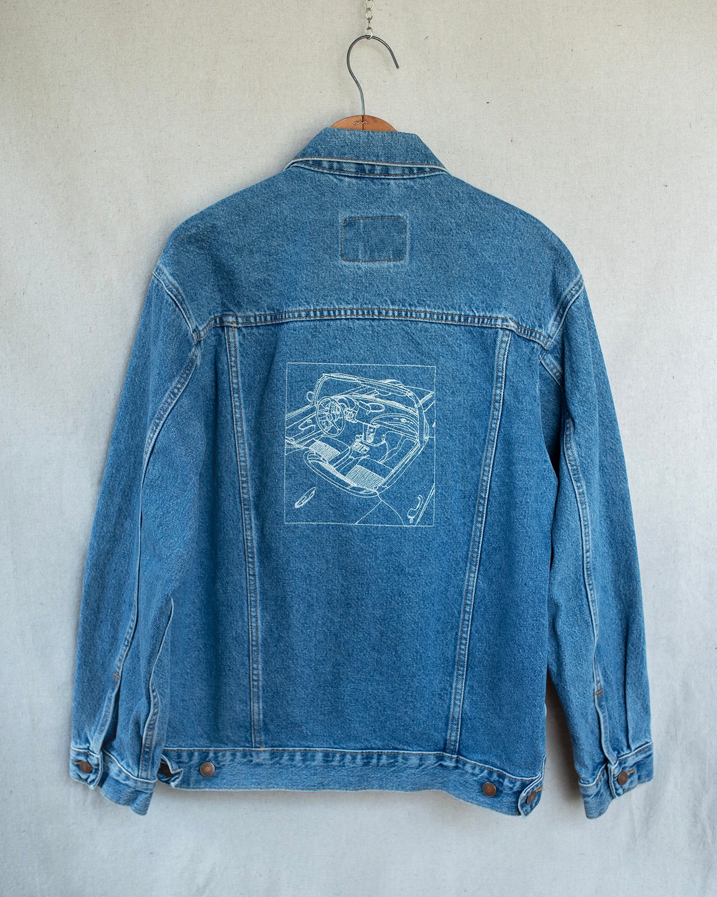 A blue denim jacket hanging on a wooden hanger against a textured off-white wall, showing the back which features an embroidery of a car.