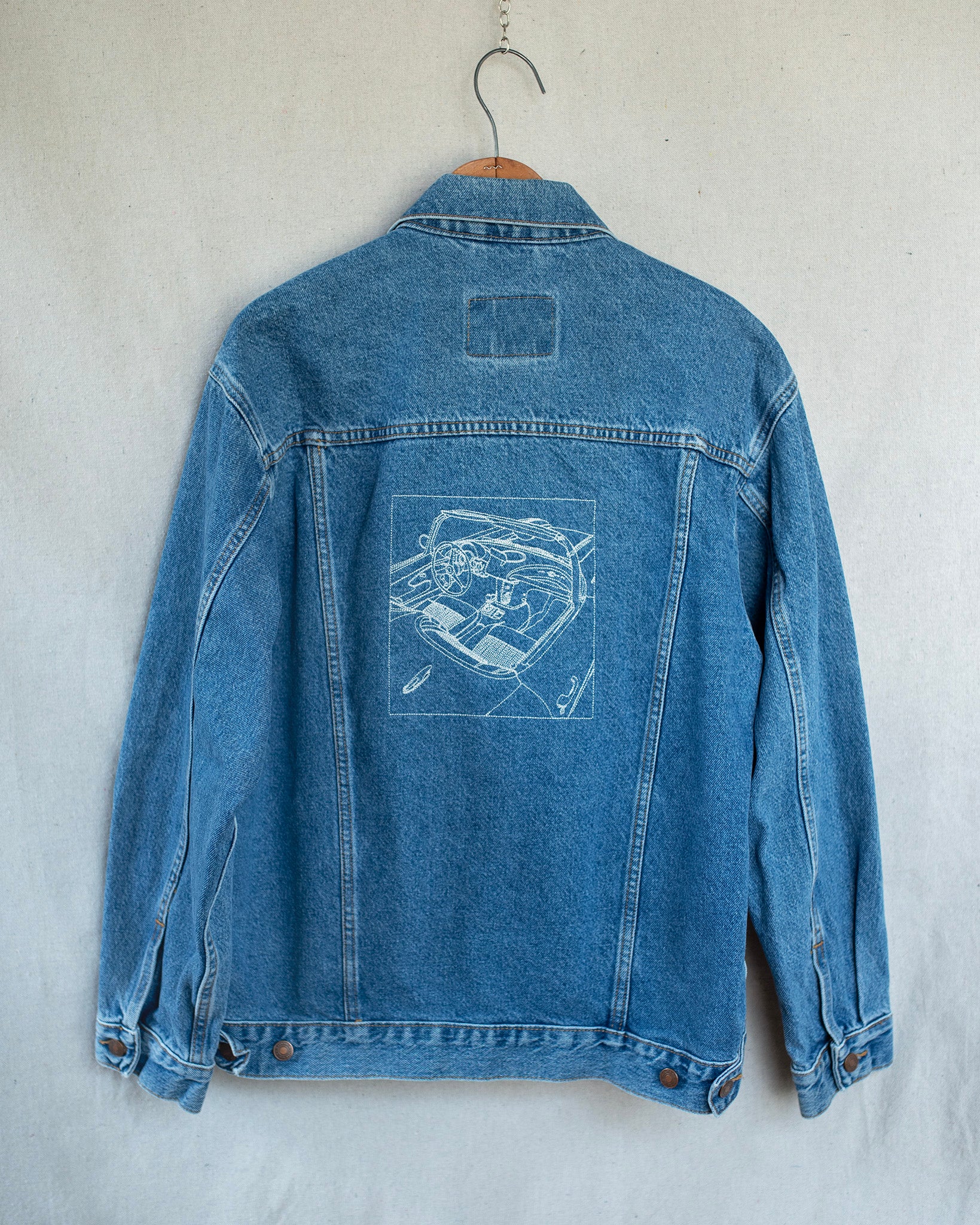 A blue denim jacket hanging on a wooden hanger against a textured off-white wall, showing the back which features an embroidery of a car.