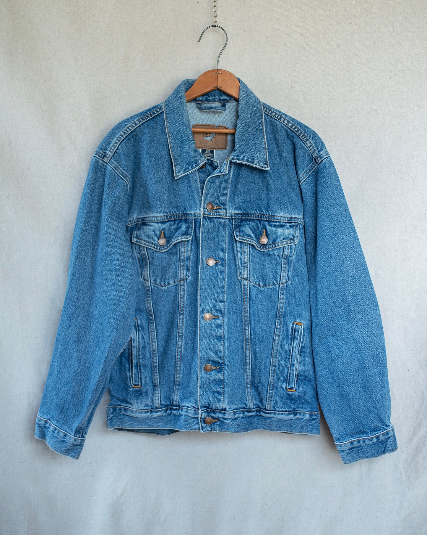 A classic blue denim jacket hanging on a wooden hanger against a light gray fabric backdrop.