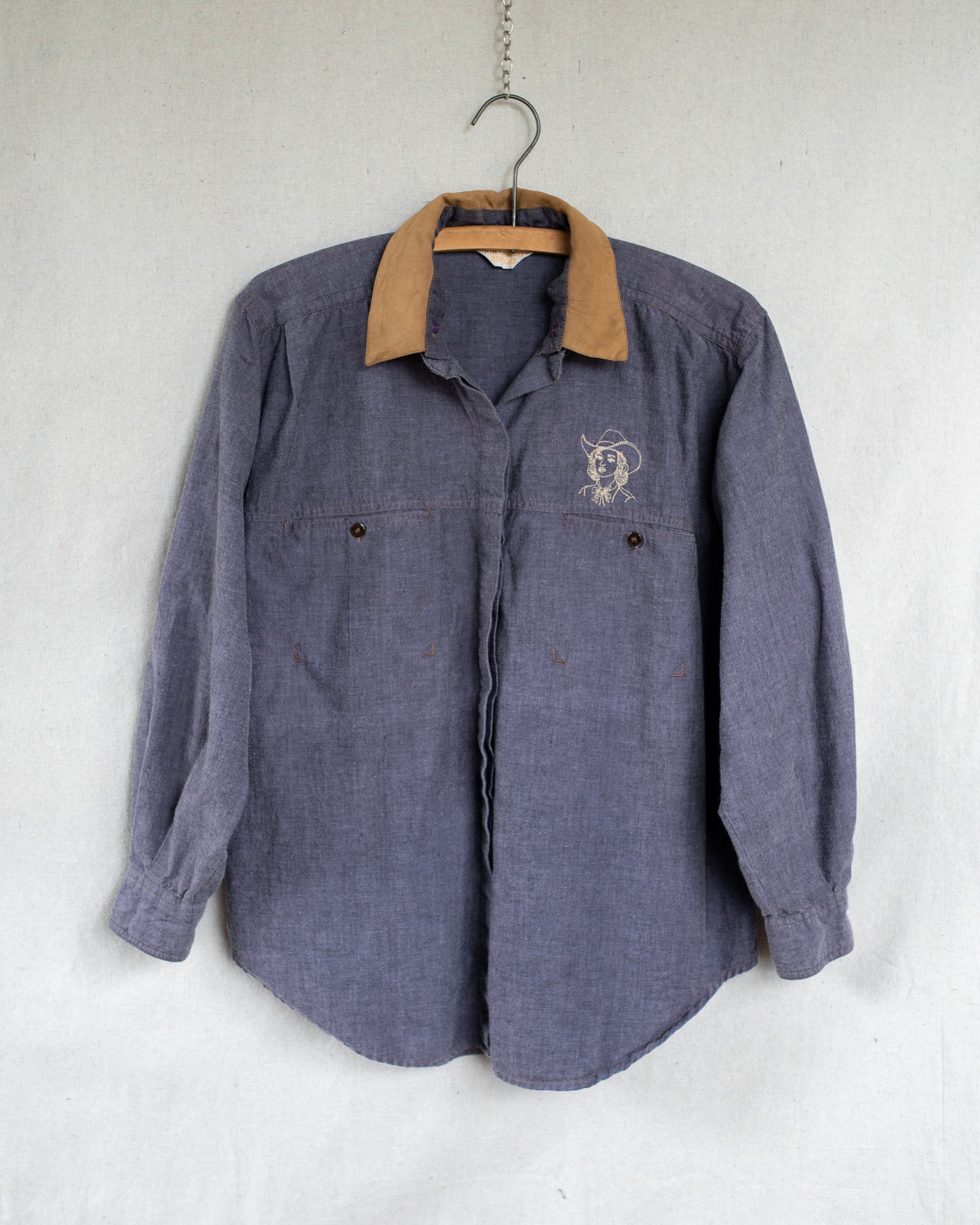 A vintage long-sleeve button-up shirt in a washed purple fabric, hanging on a wooden hanger against a light gray textured wall. It features a small embroidered cowgirl above the left chest pocket.