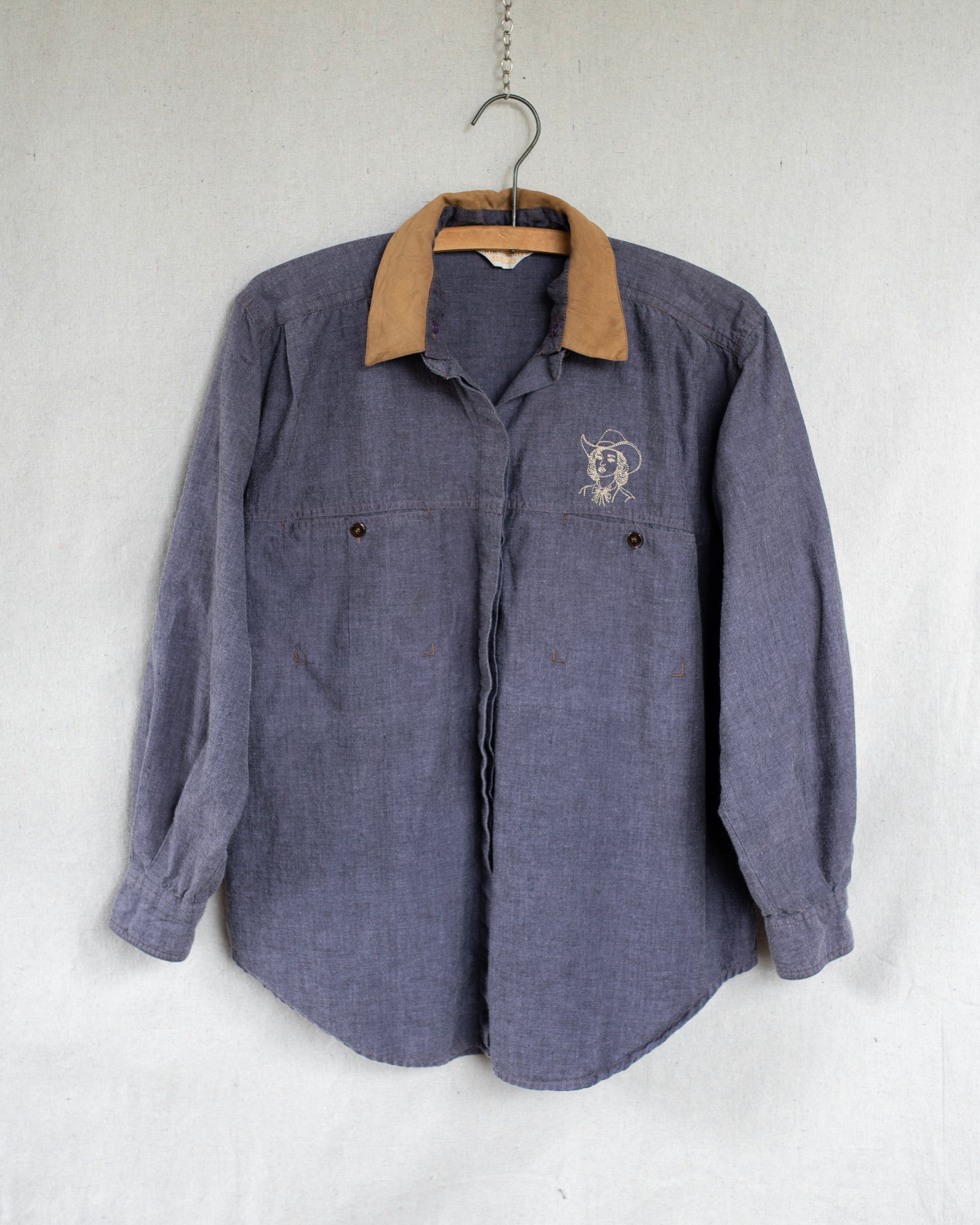 A vintage long-sleeve button-up shirt in a washed purple fabric, hanging on a wooden hanger against a light gray textured wall. It features a small embroidered cowgirl above the left chest pocket.