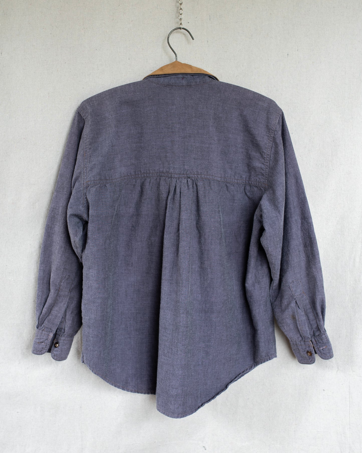 The back of a purple long-sleeve shirt with a gathered back seam, hanging on a wooden hanger against a textured off-white wall.