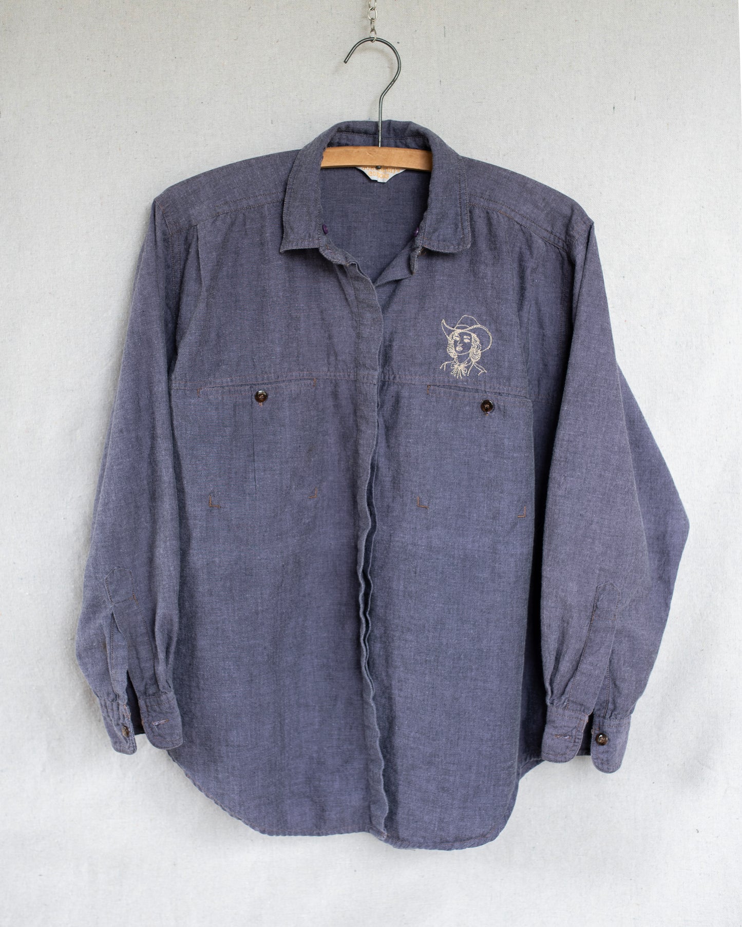 A vintage long-sleeve button-up shirt in a washed purple fabric, hanging on a wooden hanger against a light gray textured wall. It features a small embroidered cowgirl above the left chest pocket.