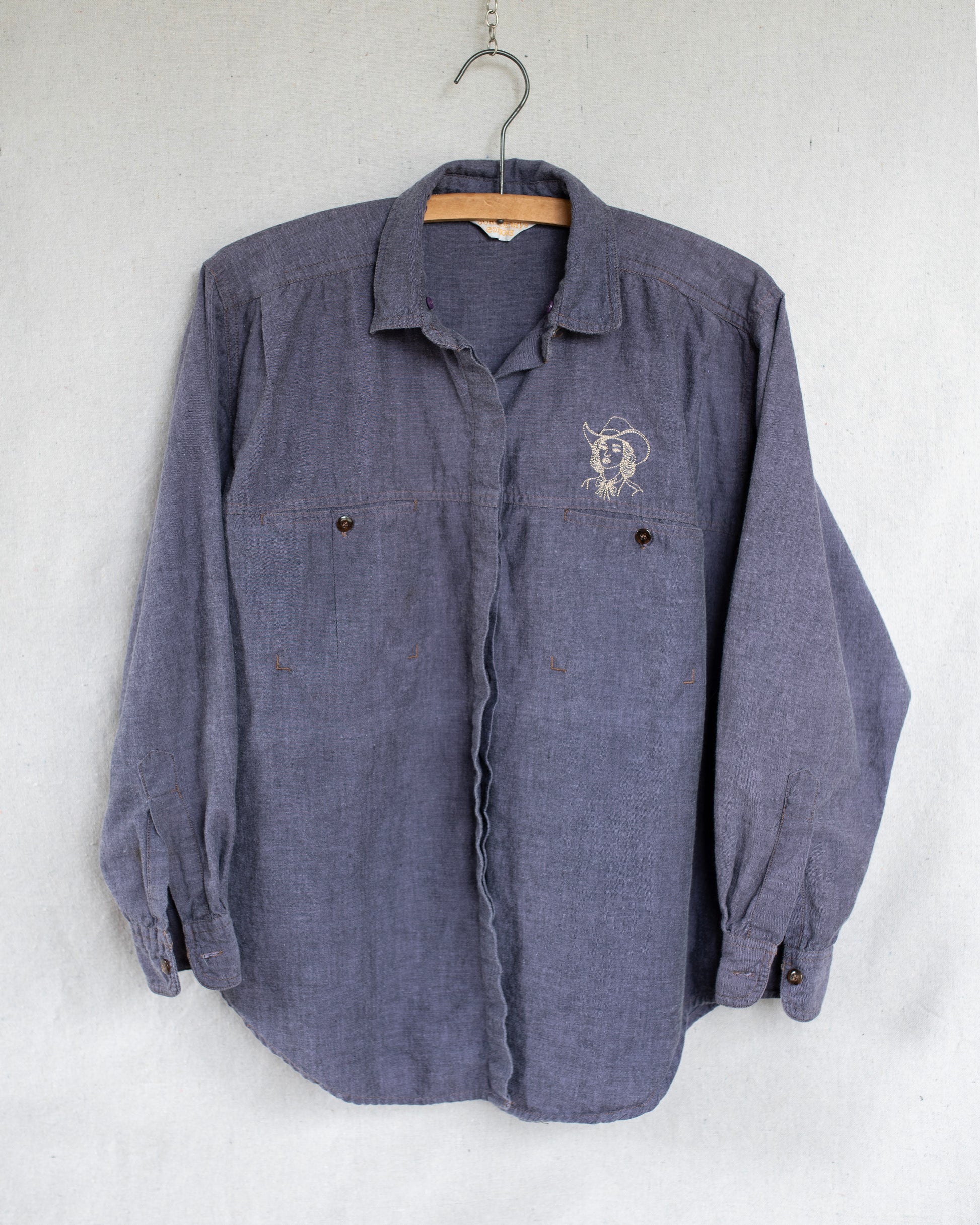 A vintage long-sleeve button-up shirt in a washed purple fabric, hanging on a wooden hanger against a light gray textured wall. It features a small embroidered cowgirl above the left chest pocket.