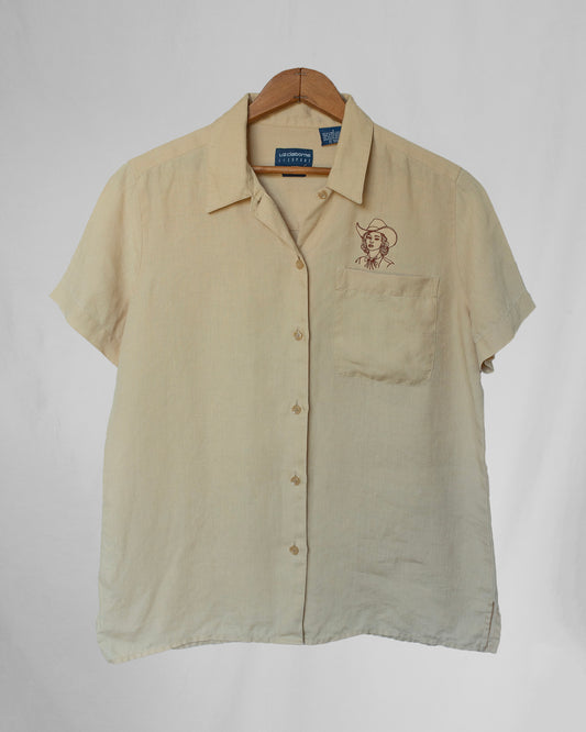 Pale yellow shirt with cowgirl embroidery above chest pocket, hanging in front of a canvas tarp