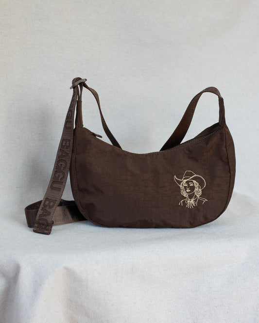 A dark brown crescent-shaped shoulder bag with a matching adjustable strap, featuring a light beige embroidered cowboy logo on the front and 'BAGGU' text on the strap, set against a light gray fabric backdrop.