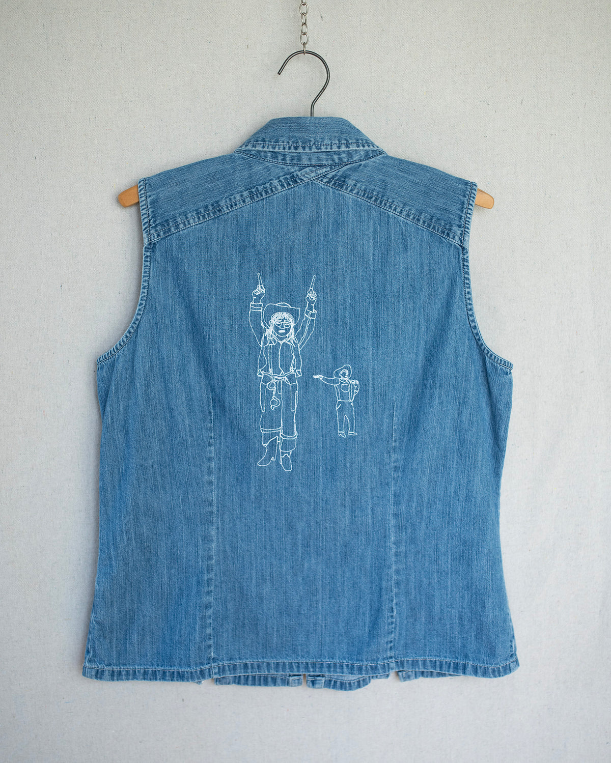 Denim top hanging from a wooden hanger, showing the back which has an embroidery of a cowgirl and big tex.