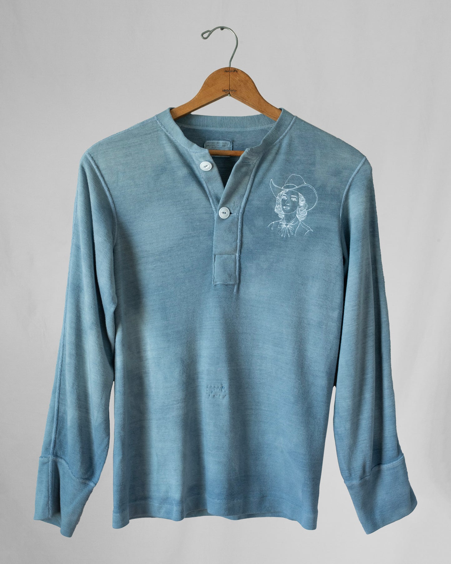 Blue long-sleeve shirt with a cowgirl embroidery on a hanger against a gray background.