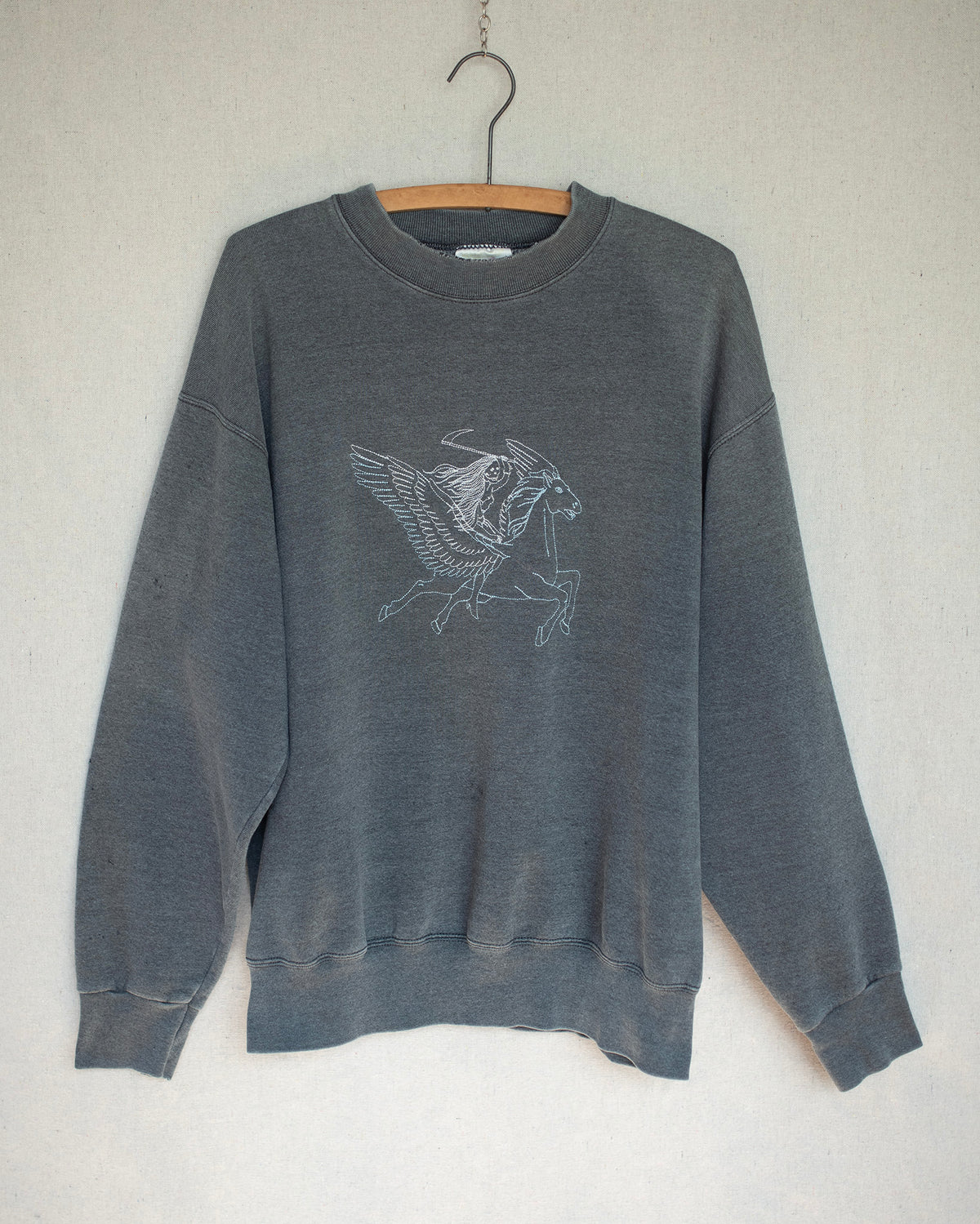Dark grey crewneck sweatshirt with a two tone embroidery on front hanging on a wooden hanger.