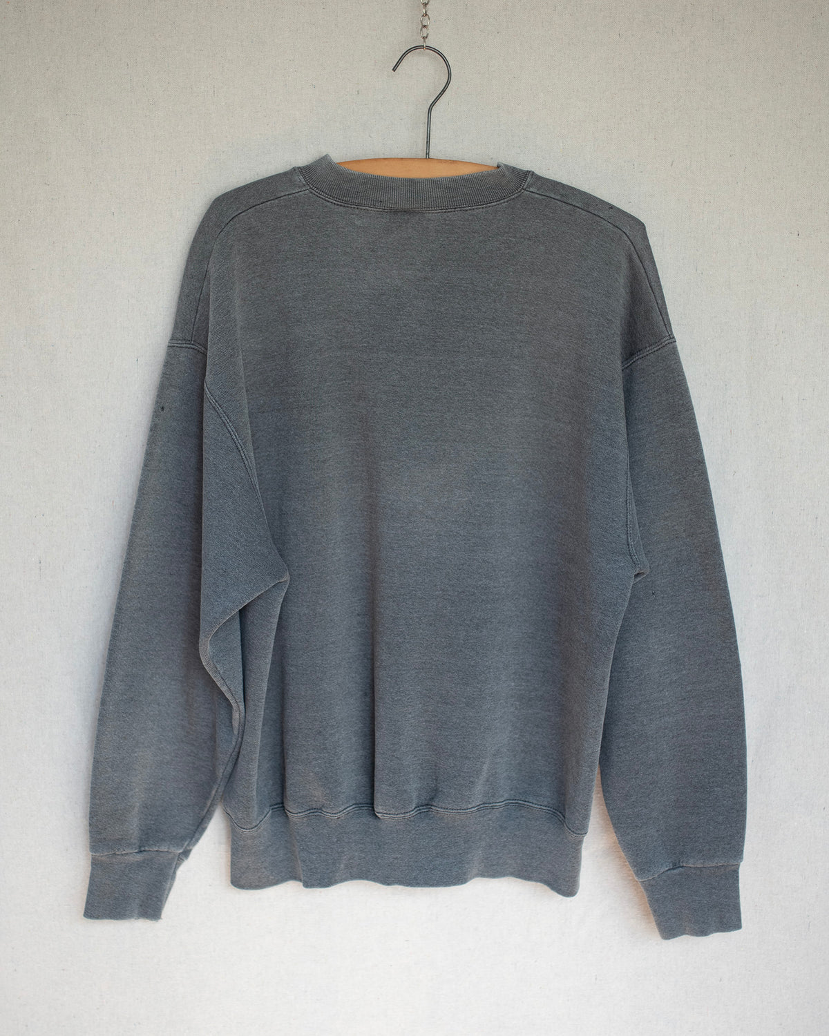 A faded dark gray crewneck sweatshirt hanging on a wooden hanger against a textured off-white wall.