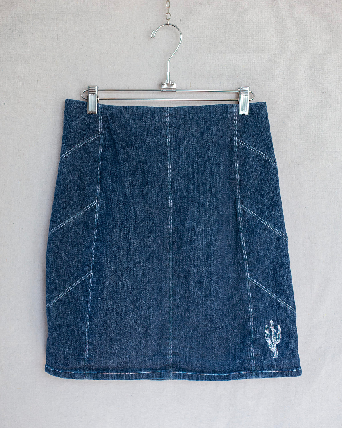 Denim skirt hanging on a metal hanger with a small cactus embroidery located in the corner.
