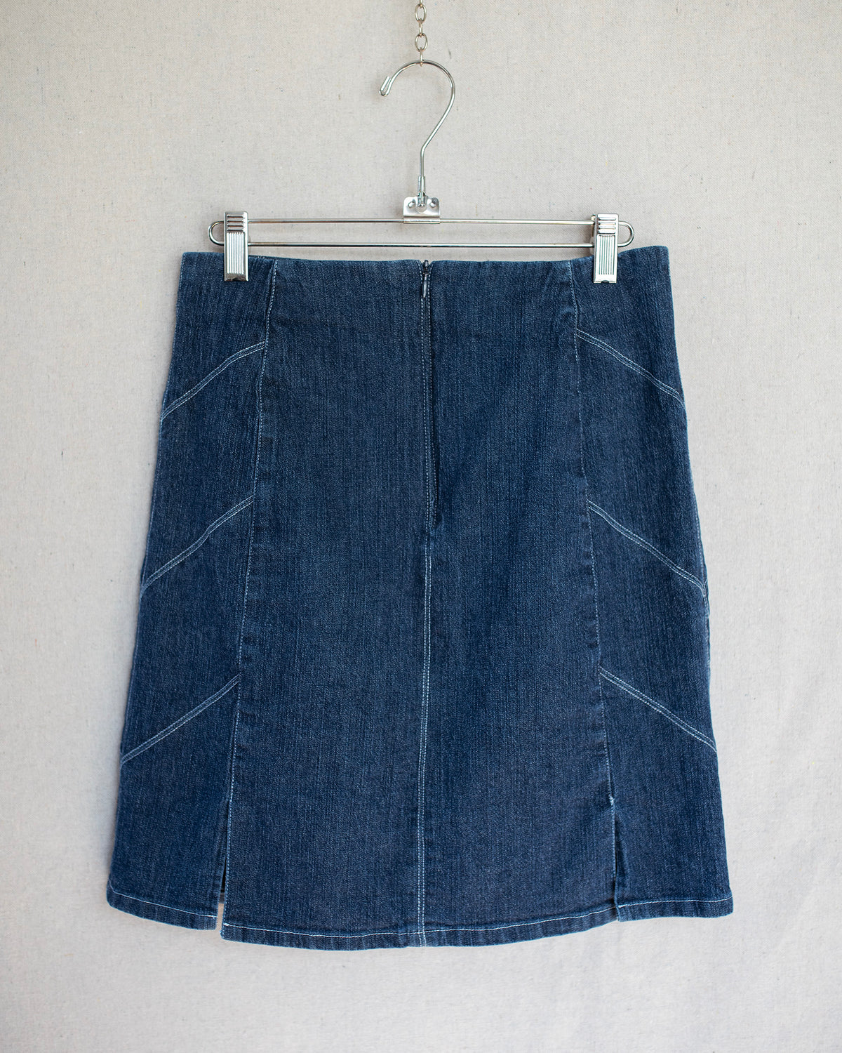 A dark blue denim mini skirt hanging on a metal hanger against a light gray fabric background.