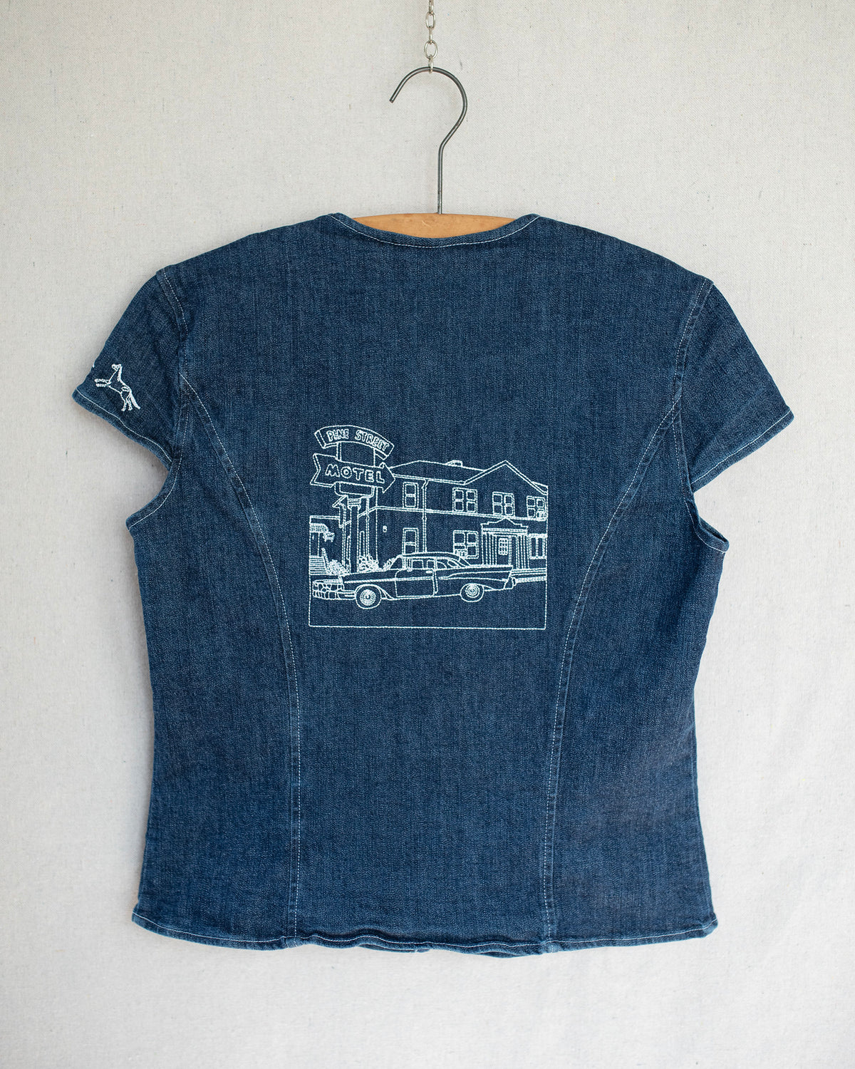Denim top hanging on a wooden hanger, showing the back with embroidery details.