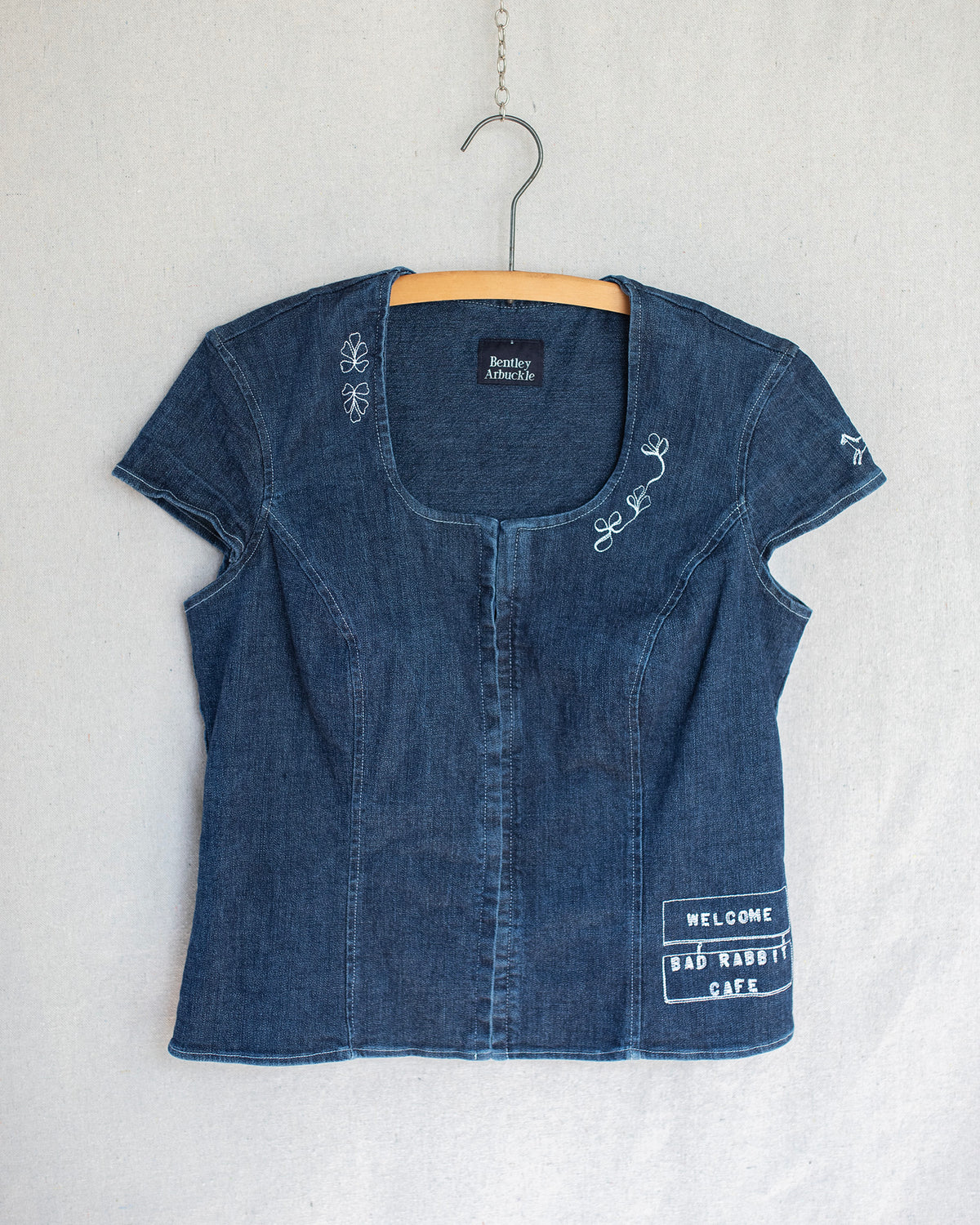 A blue denim short-sleeve top with white embroidered details, hanging on a wooden hanger against a textured off-white wall.