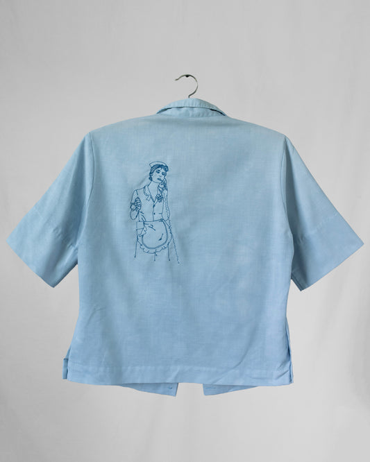 Light blue shirt with an embroidered design on back displayed on a hanger against a gray background