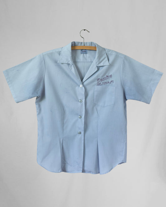 Light blue short-sleeve shirt with embroidery above chest pocket on a hanger against a gray background