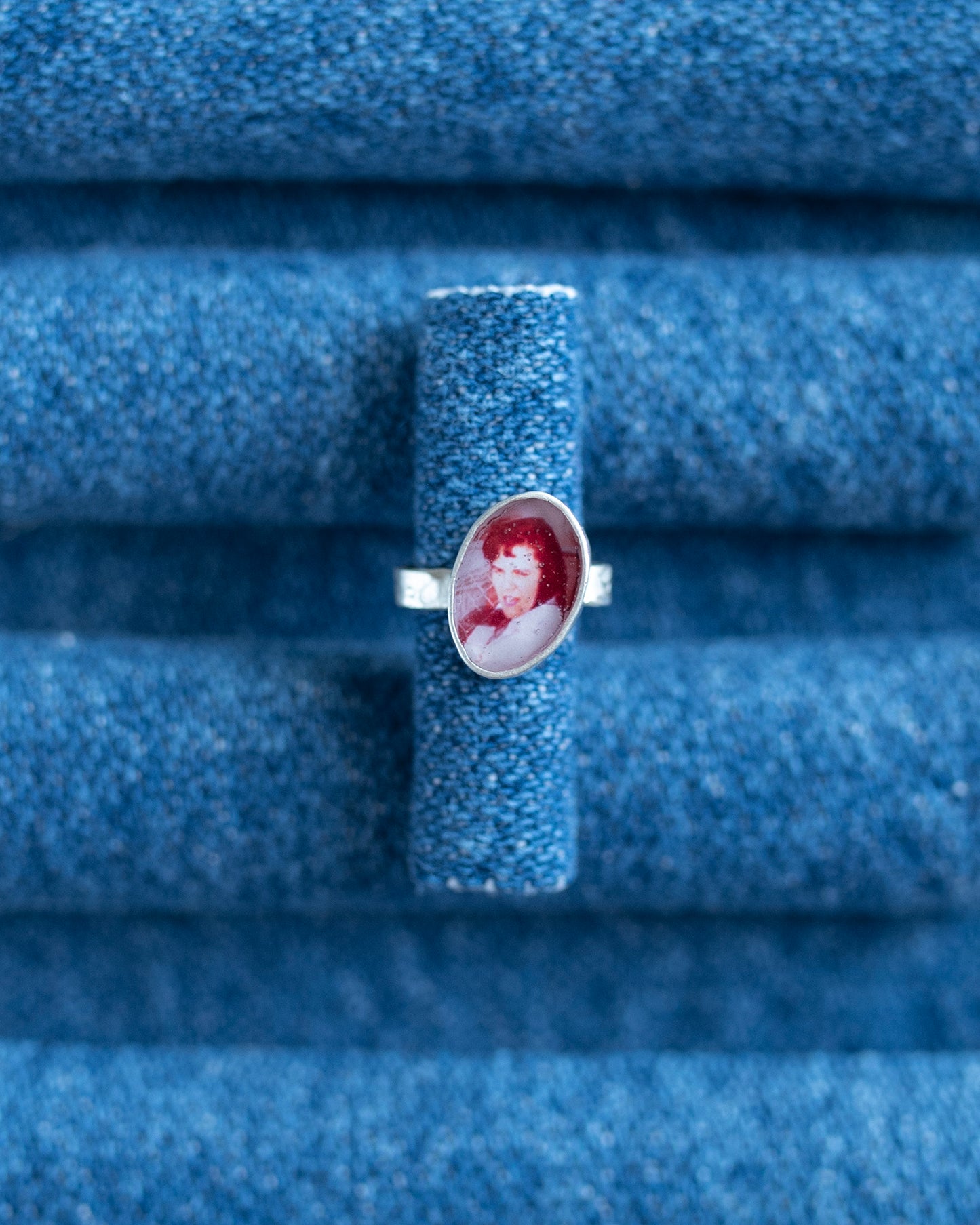 A silver band ear cuff featuring an oval portrait cabochon with red hair and a white face, displayed on a textured blue ring stand.