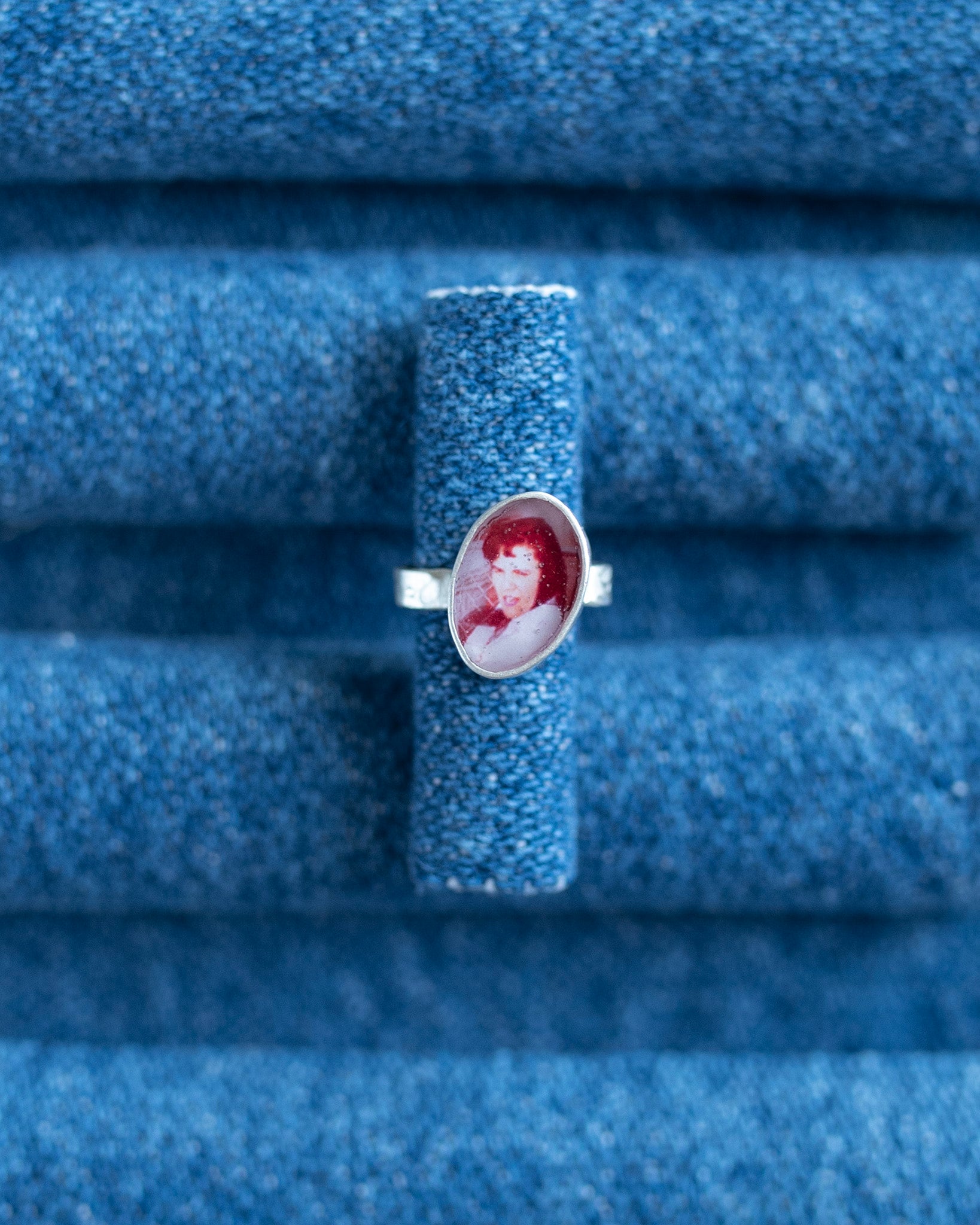 A silver band ear cuff featuring an oval portrait cabochon with red hair and a white face, displayed on a textured blue ring stand.