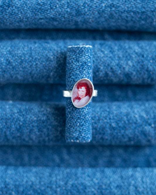 A silver band ear cuff featuring an oval portrait cabochon with red hair and a white face, displayed on a textured blue ring stand.