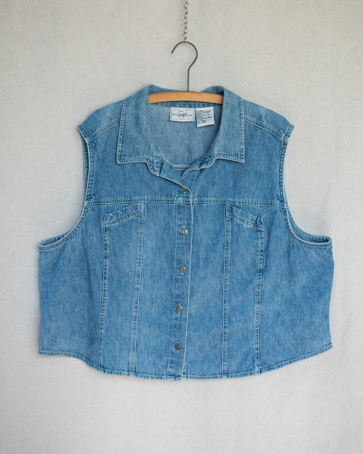 A light blue denim top with a pointed collar and button-front closure, hanging on a wooden hanger against a light gray textured wall.