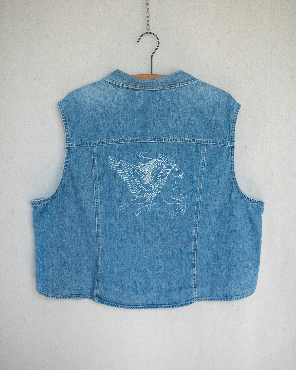 A light blue denim top hanging on a wooden hanger against a light gray wall, featuring a white embroidered Pegasus design on the back.