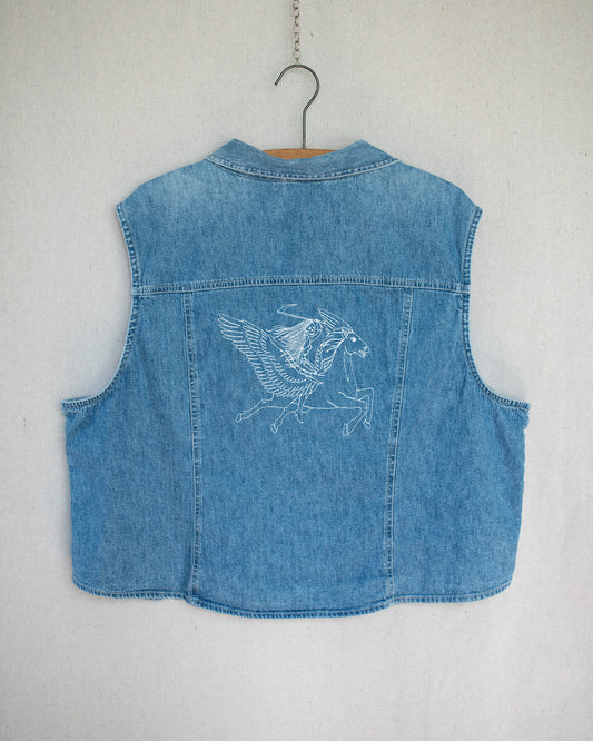 A light blue denim top hanging on a wooden hanger against a light gray wall, featuring a white embroidered Pegasus design on the back.