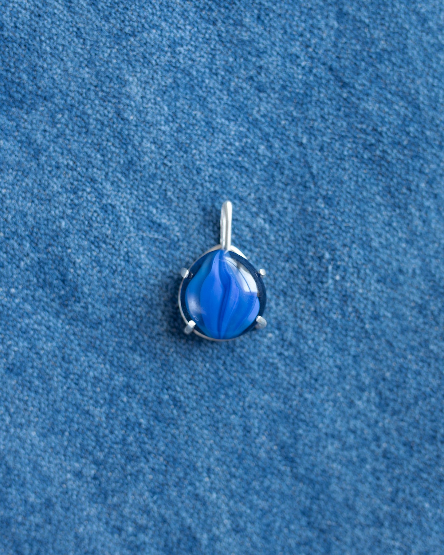 A small silver teardrop pendant with a vibrant blue marbled or swirled inlay, resting on a textured blue fabric background.
