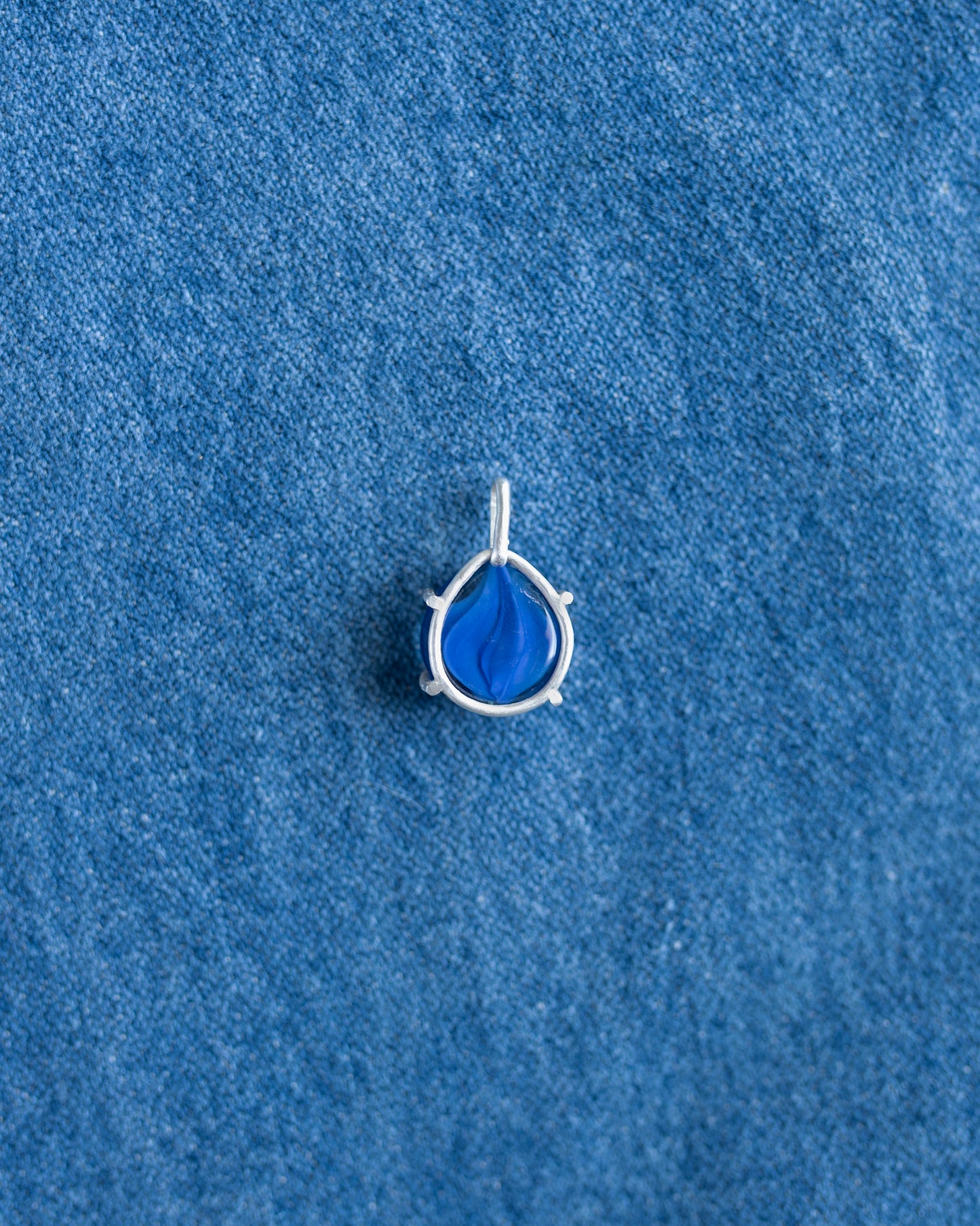 A small silver teardrop pendant with a vibrant blue marbled gemstone, displayed on a textured blue fabric background.