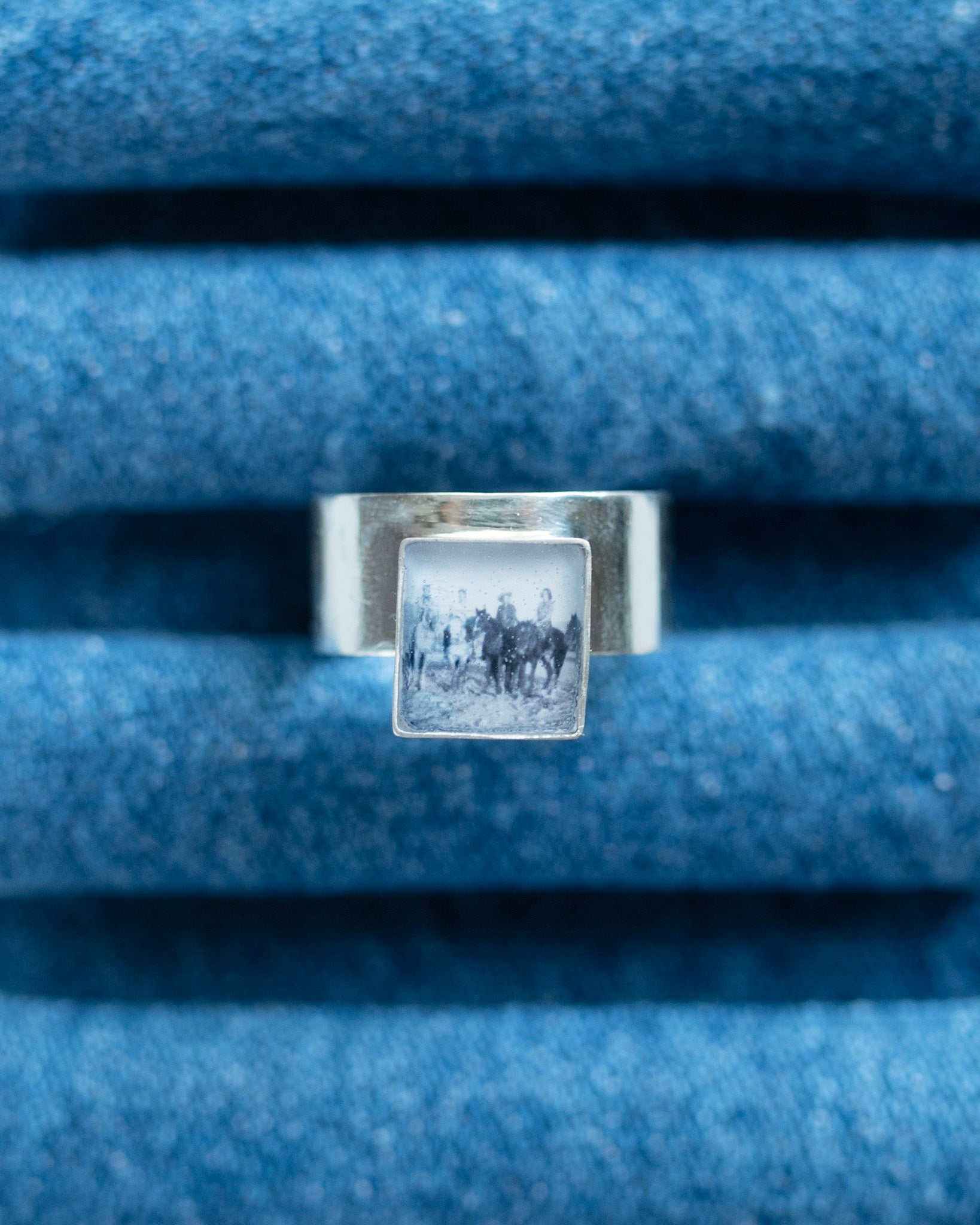 A silver ring with a square bezel containing a black-and-white vintage photograph of a horse riders, displayed on a textured blue denim ring stand.