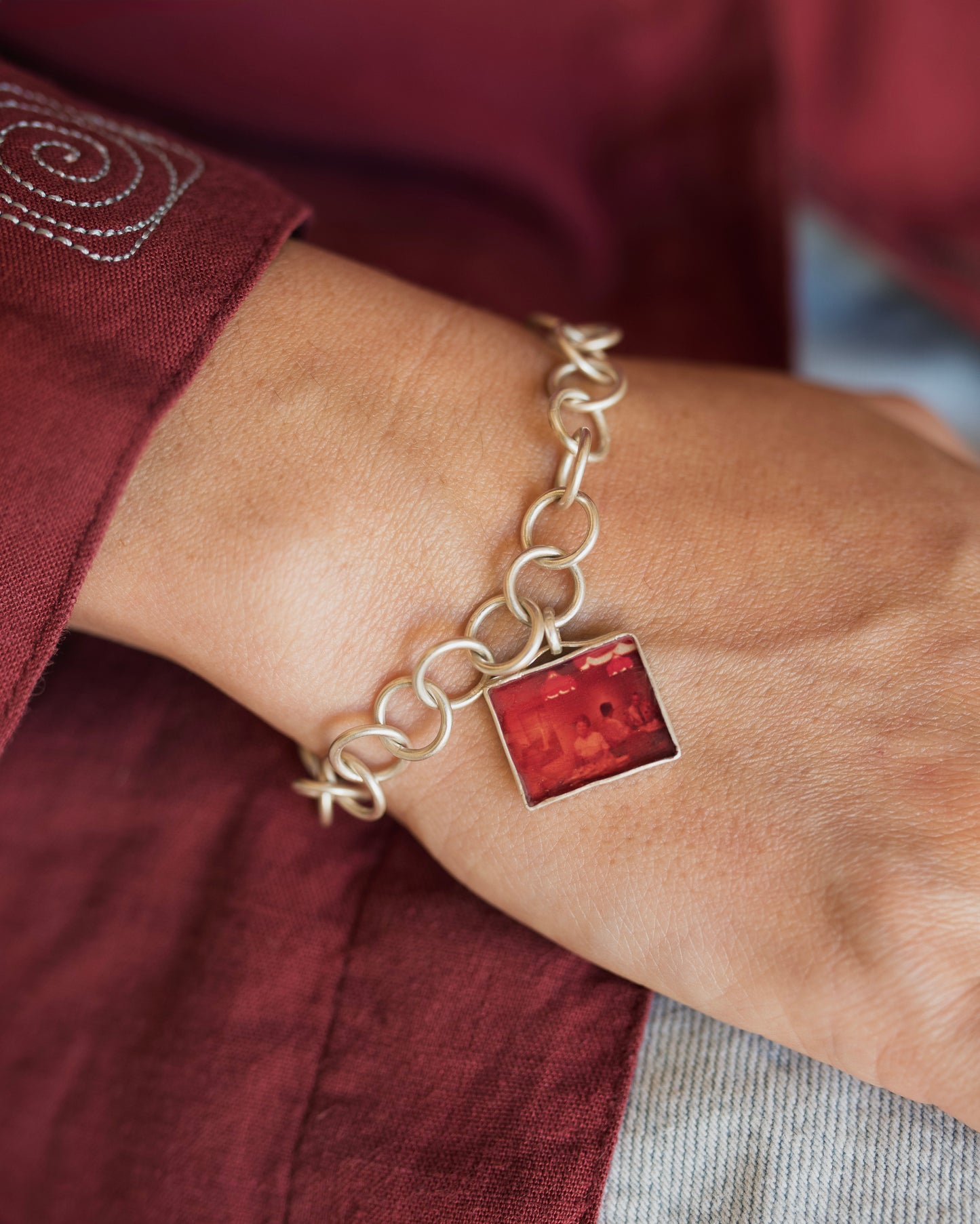 A close-up of a person's wrist wearing a gold chain bracelet with a square red stone pendant, set against a background of a burgundy shirt.