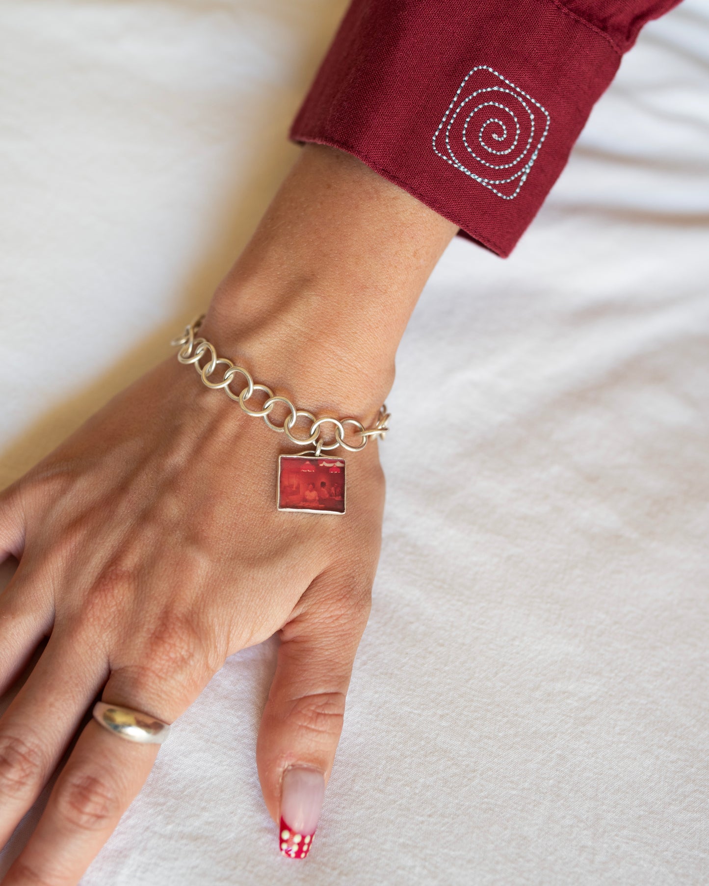 A close-up of a person's hand wearing a silver chain bracelet with a square red textured charm, resting on a white fabric background.